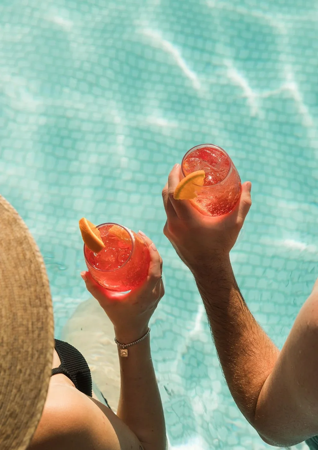 Two people relaxing in a swimming pool, holding glasses of pink drinks with ice and orange slices.
