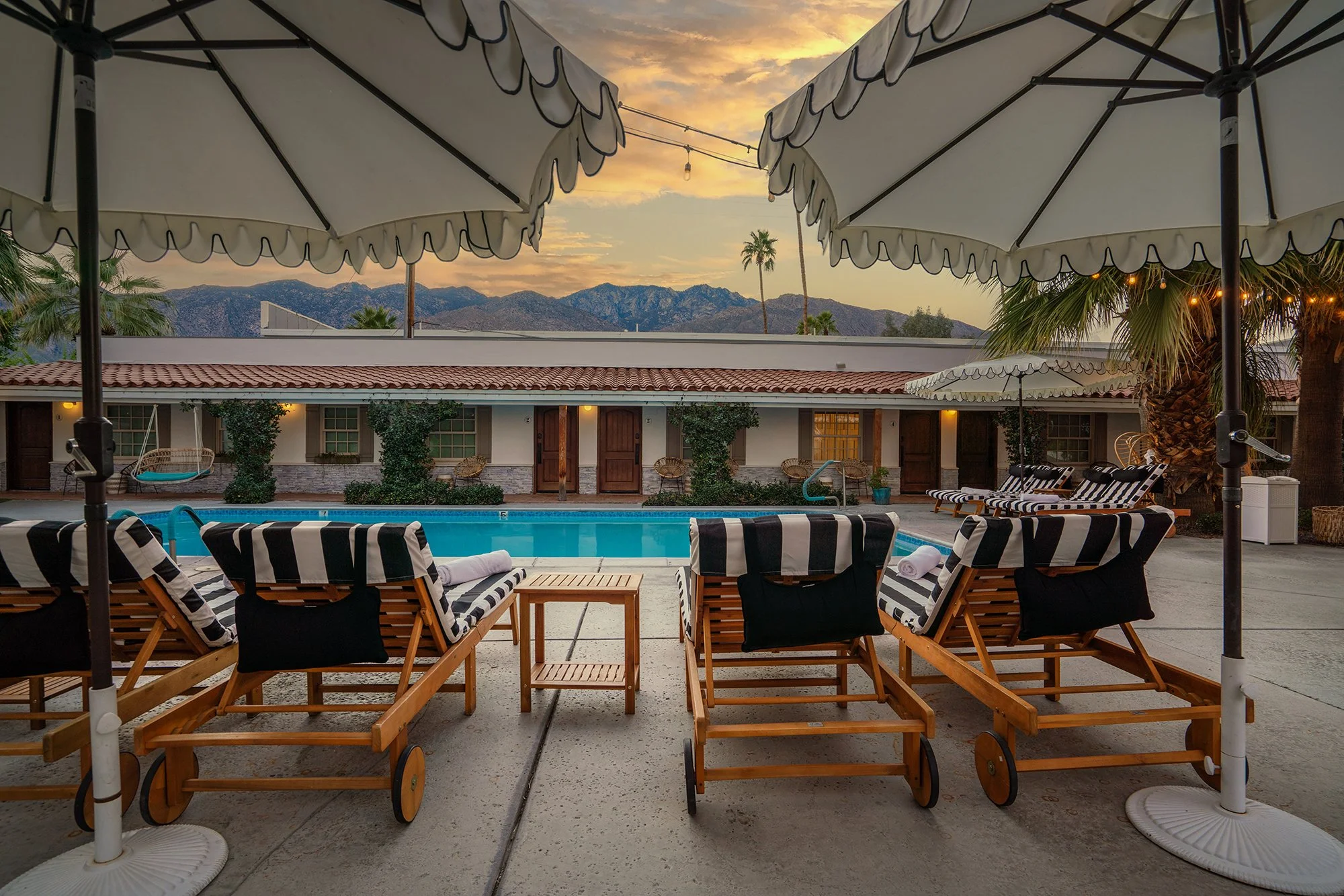 Swimming pool surrounded by wooden lounge chairs with black and white striped cushions and umbrellas, set against a backdrop of mountains during sunset.