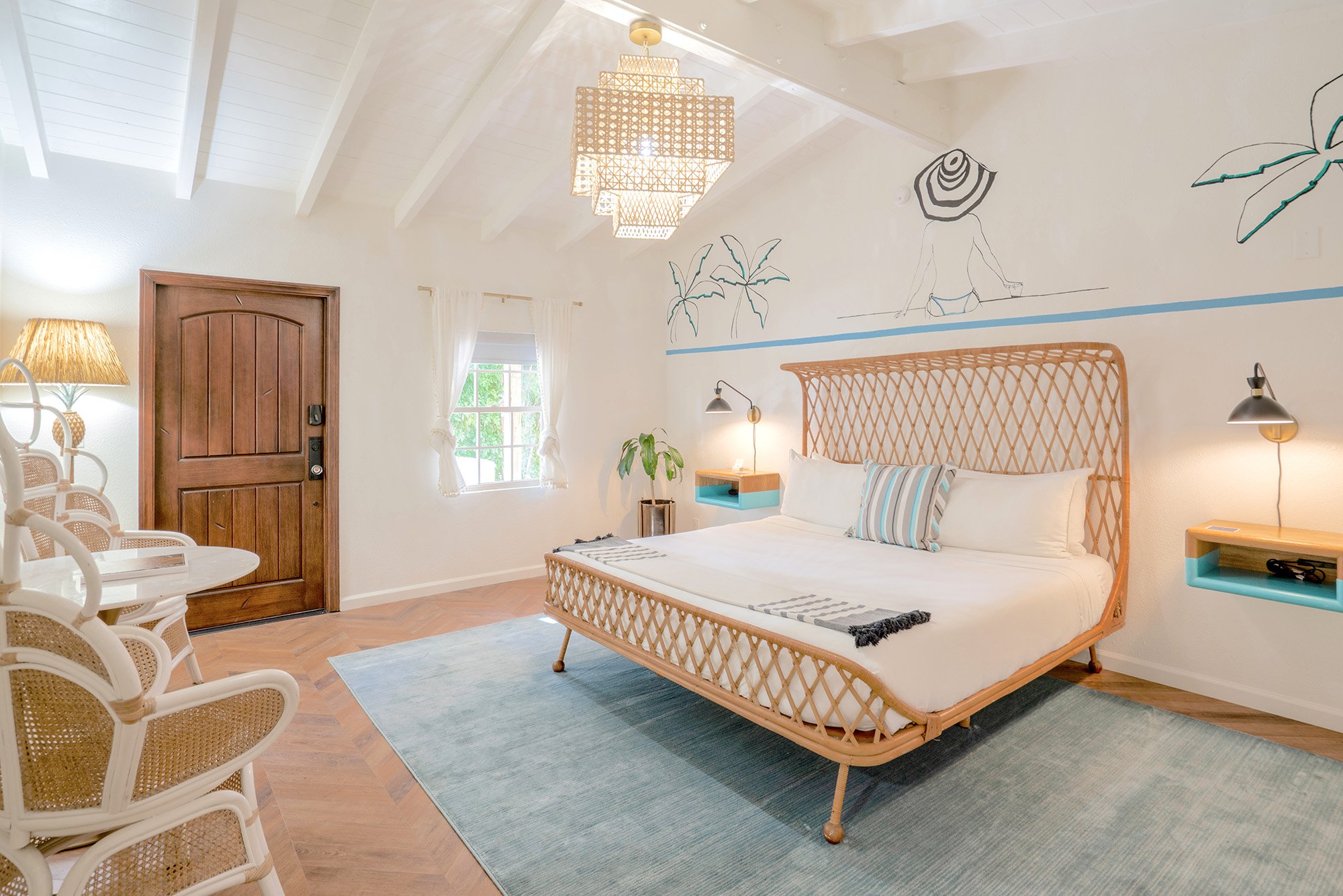 Cozy bedroom with a wooden door, white walls with beach-themed artwork, a queen-sized bed with a woven headboard, nautical-style pillows, floating side tables, a small window with white curtains, a plant near the bed, wicker chairs, and a woven chand