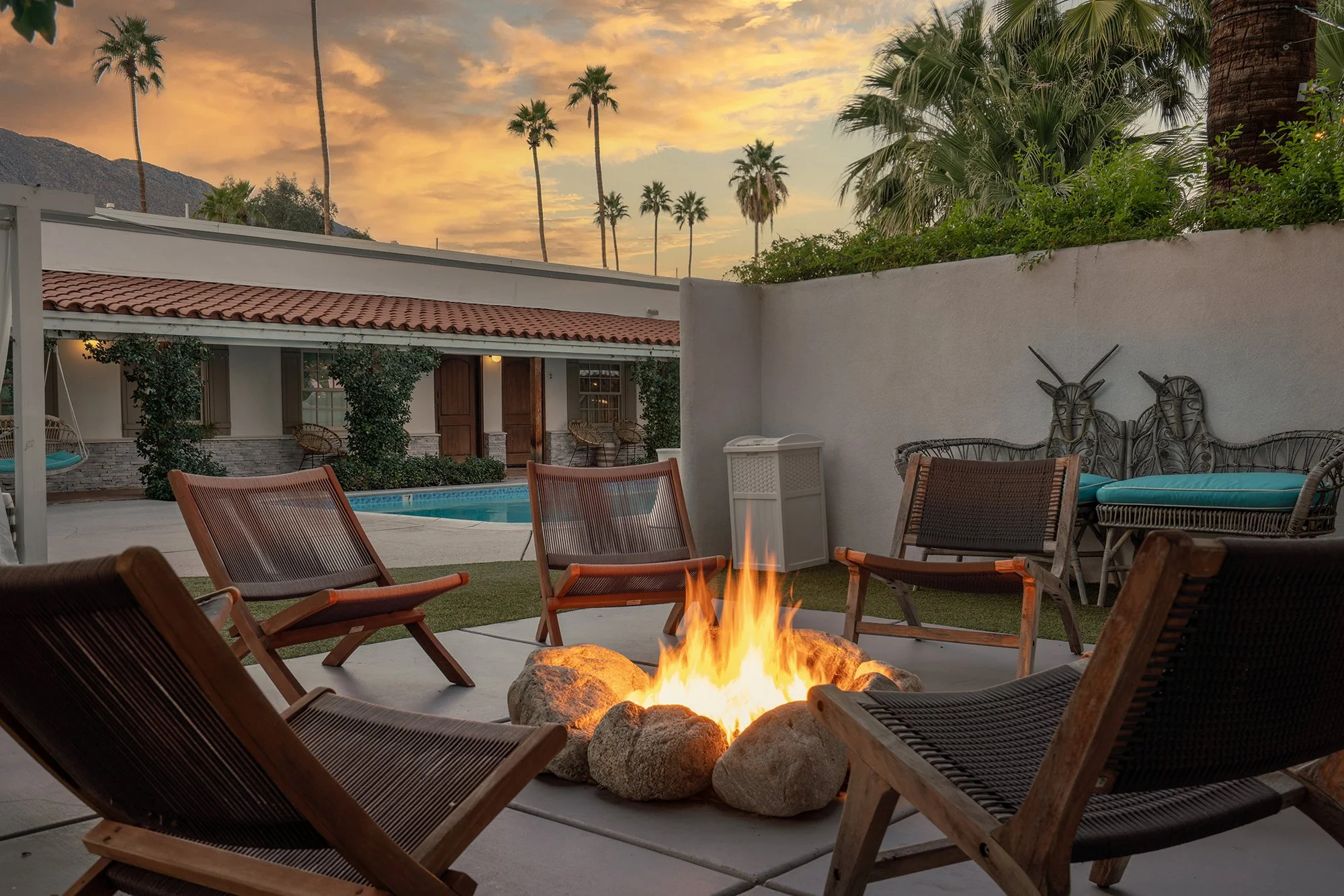 A cozy outdoor patio with a fire pit surrounded by wooden chairs, overlooking a swimming pool and a house with a red-tiled roof at sunset, with palm trees in the background.