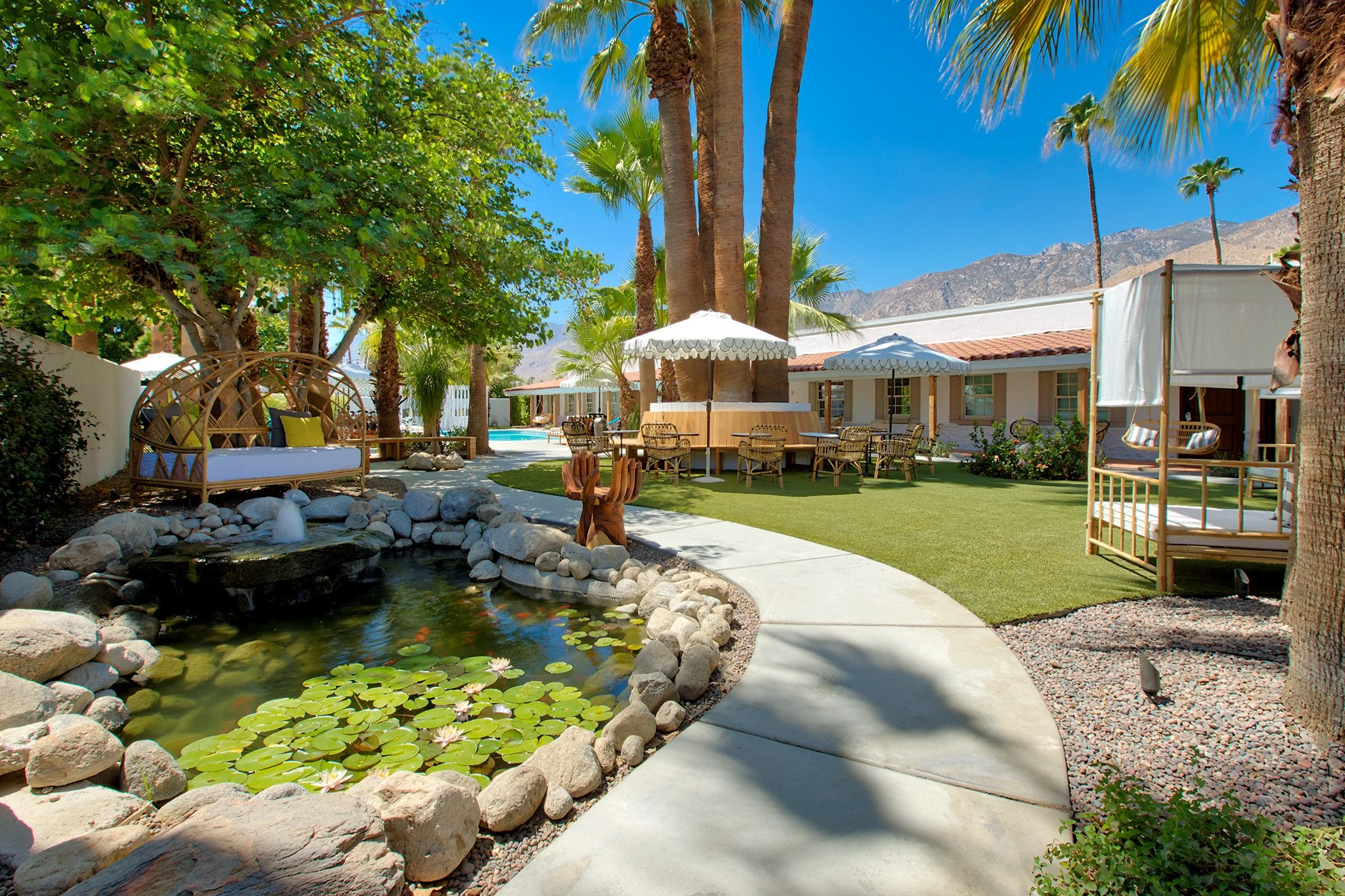 A landscaped outdoor patio area with a pond, numerous palm trees, seating areas with umbrellas, a lawn, and a mountain backdrop.