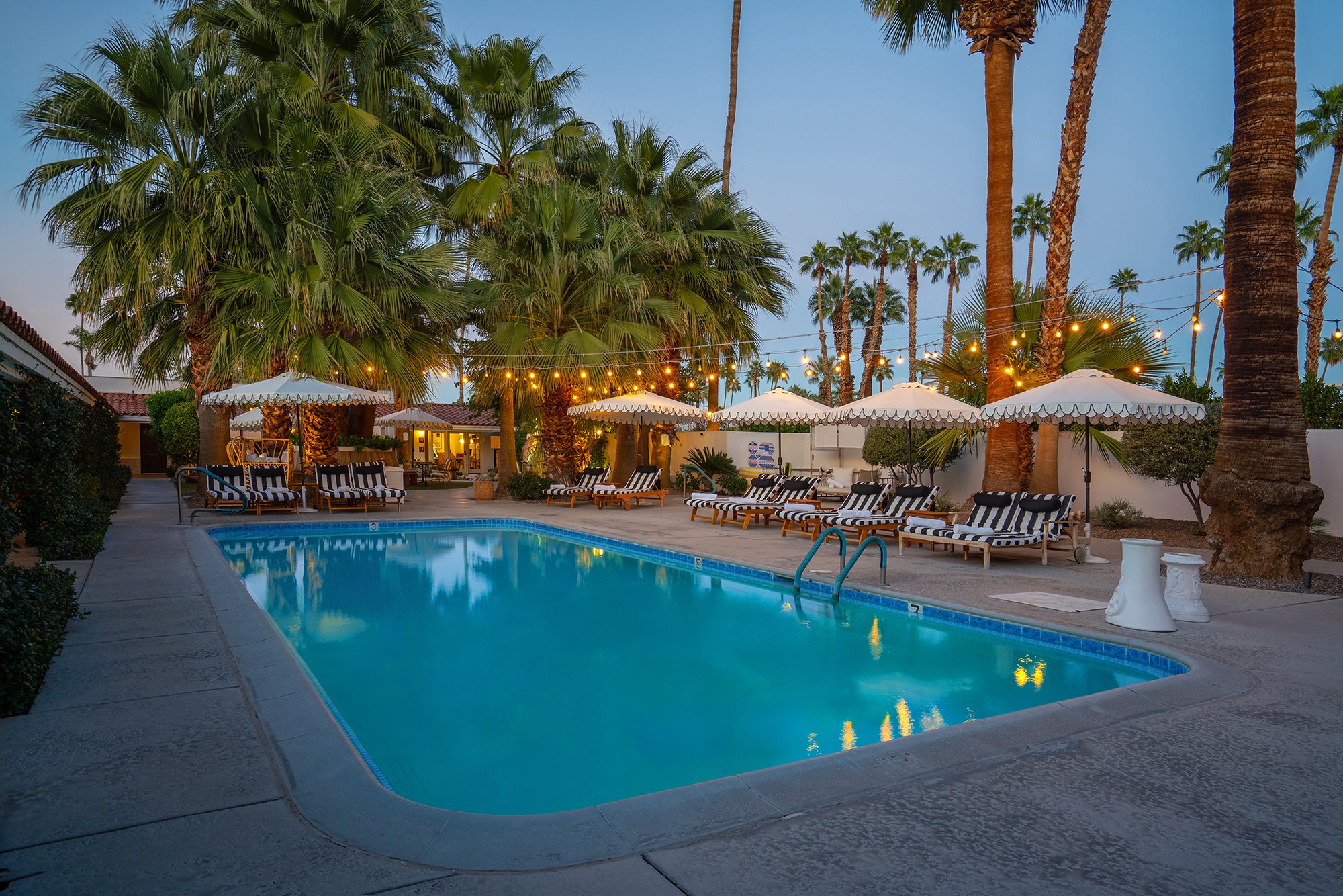 A resort swimming pool area at dusk with lounge chairs, umbrellas, string lights, tall palm trees, and a building in the background.