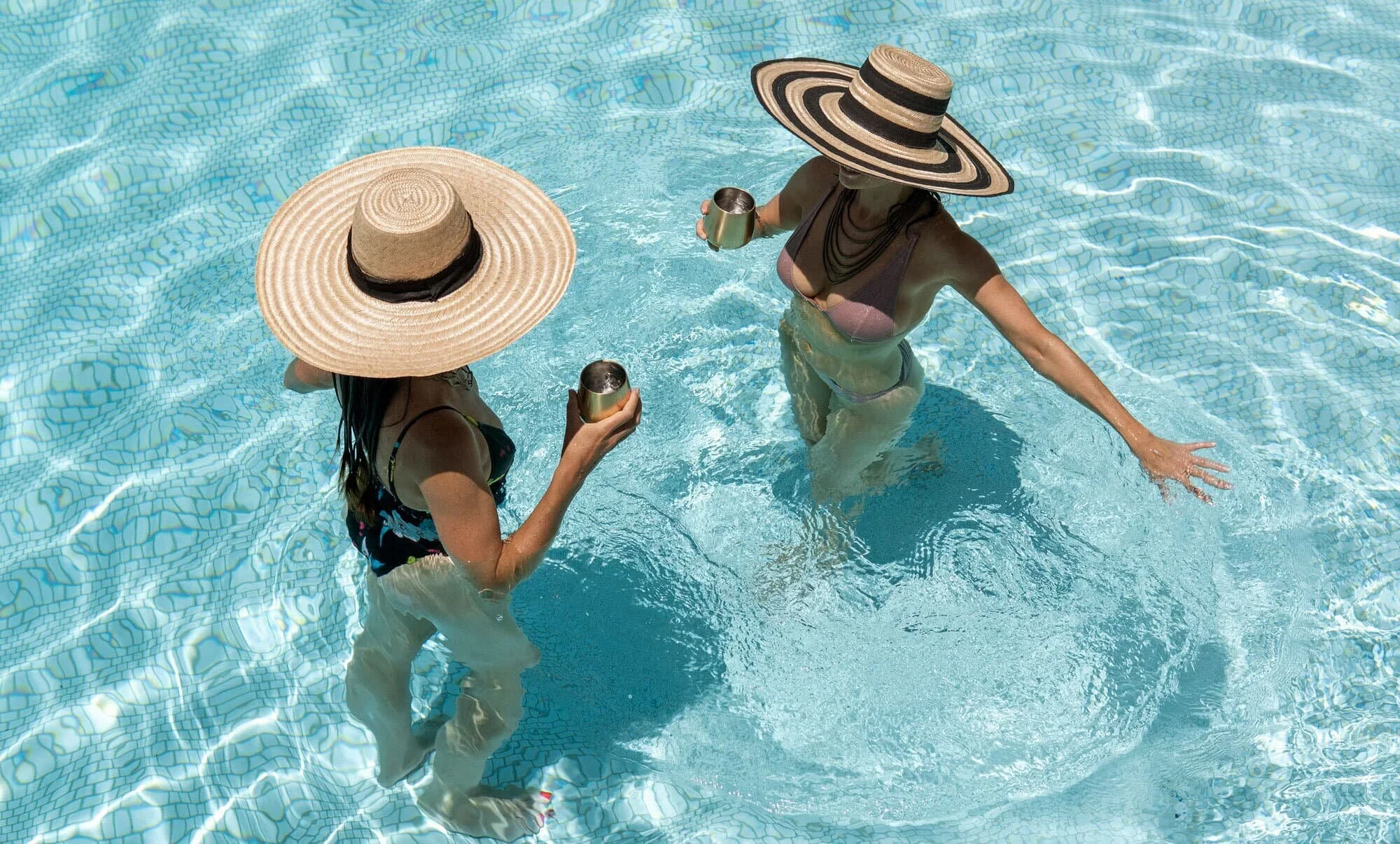 Two women in a swimming pool wearing large straw hats and holding metal cups.