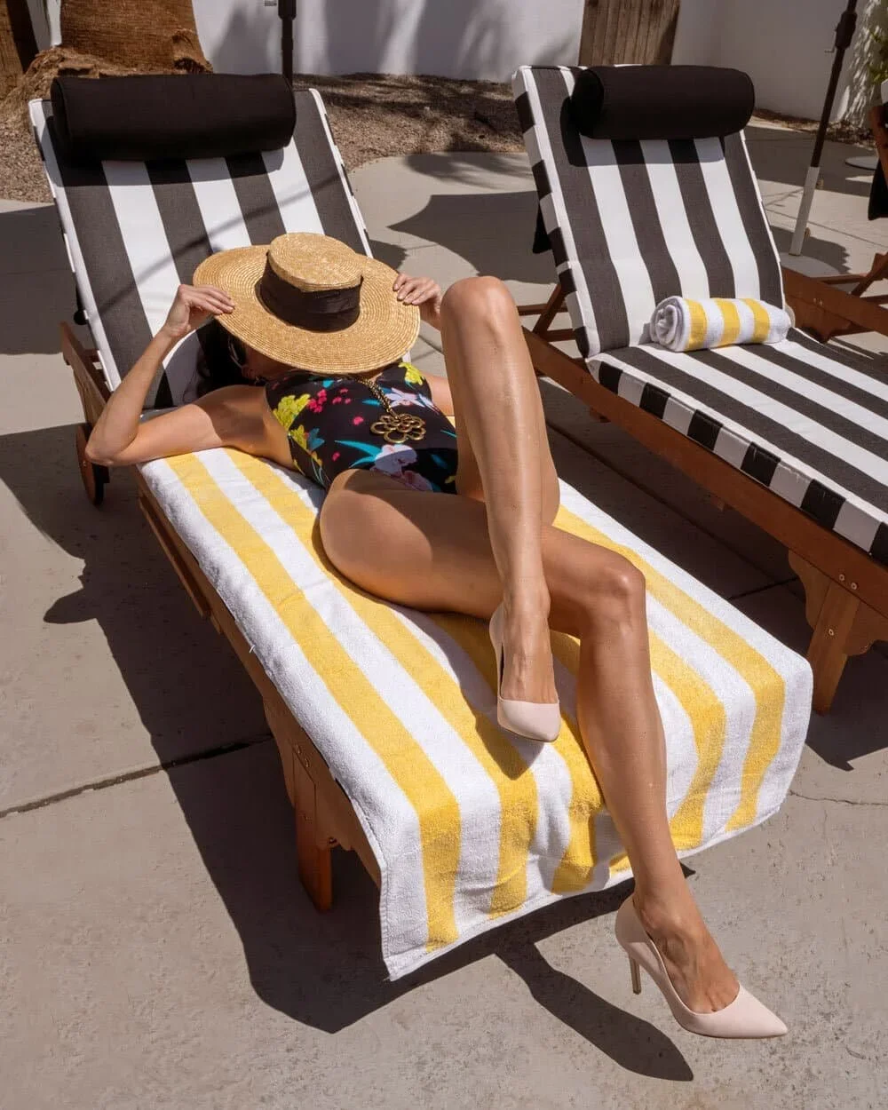Woman lounging on a poolside lounge chair with yellow and white striped towel, wearing a hat and floral swimsuit, next to another lounge chair with black and white stripes and a rolled towel.