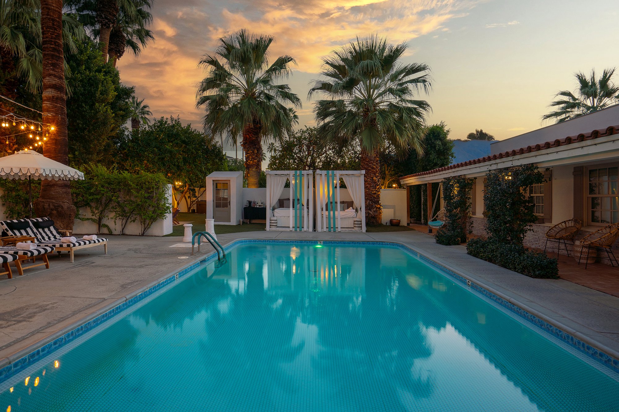 A serene backyard scene during sunset featuring a swimming pool surrounded by palm trees, outdoor furniture, and a shaded cabana with curtains.