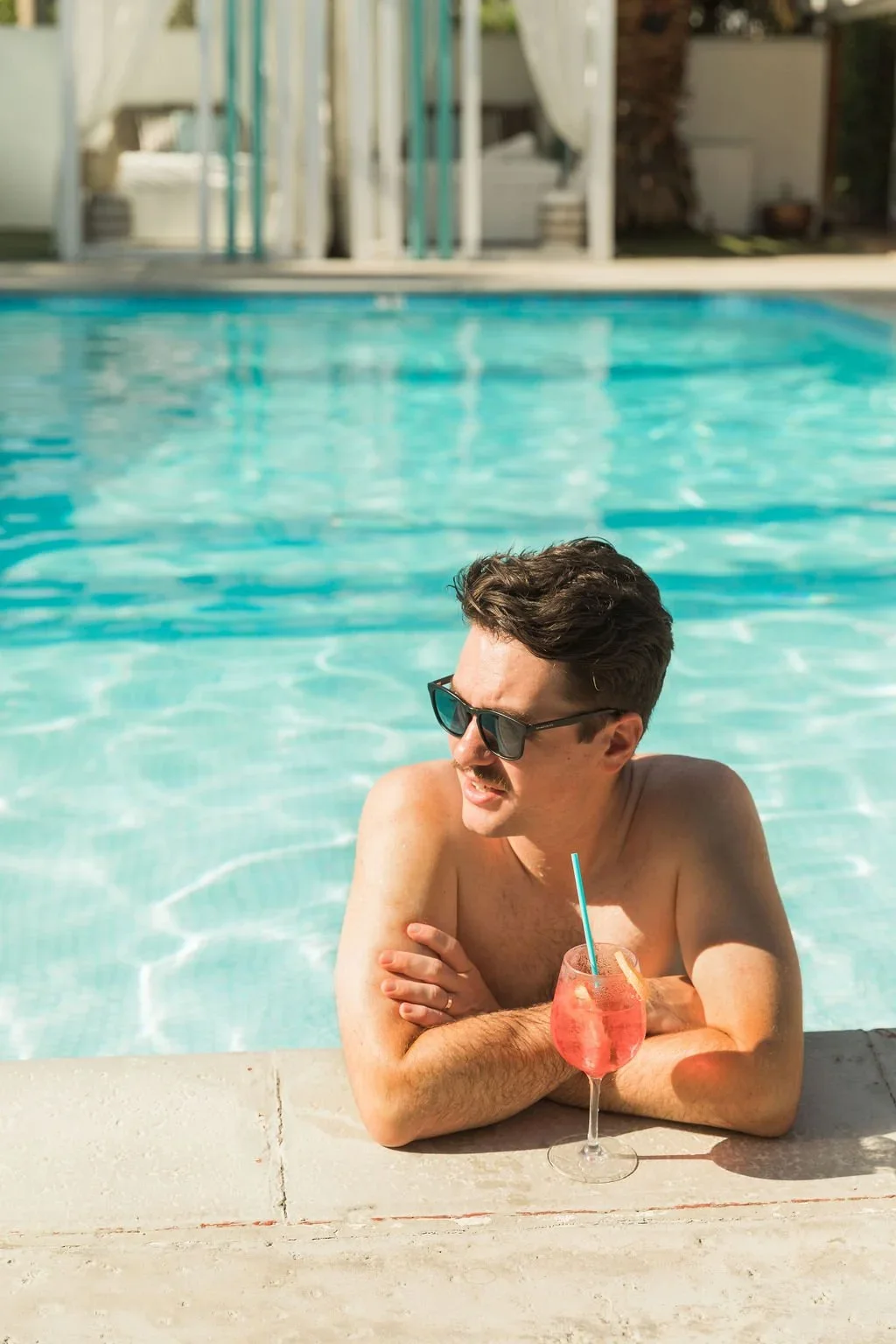 A man with sunglasses relaxing at a swimming pool with a pink cocktail in a wine glass.