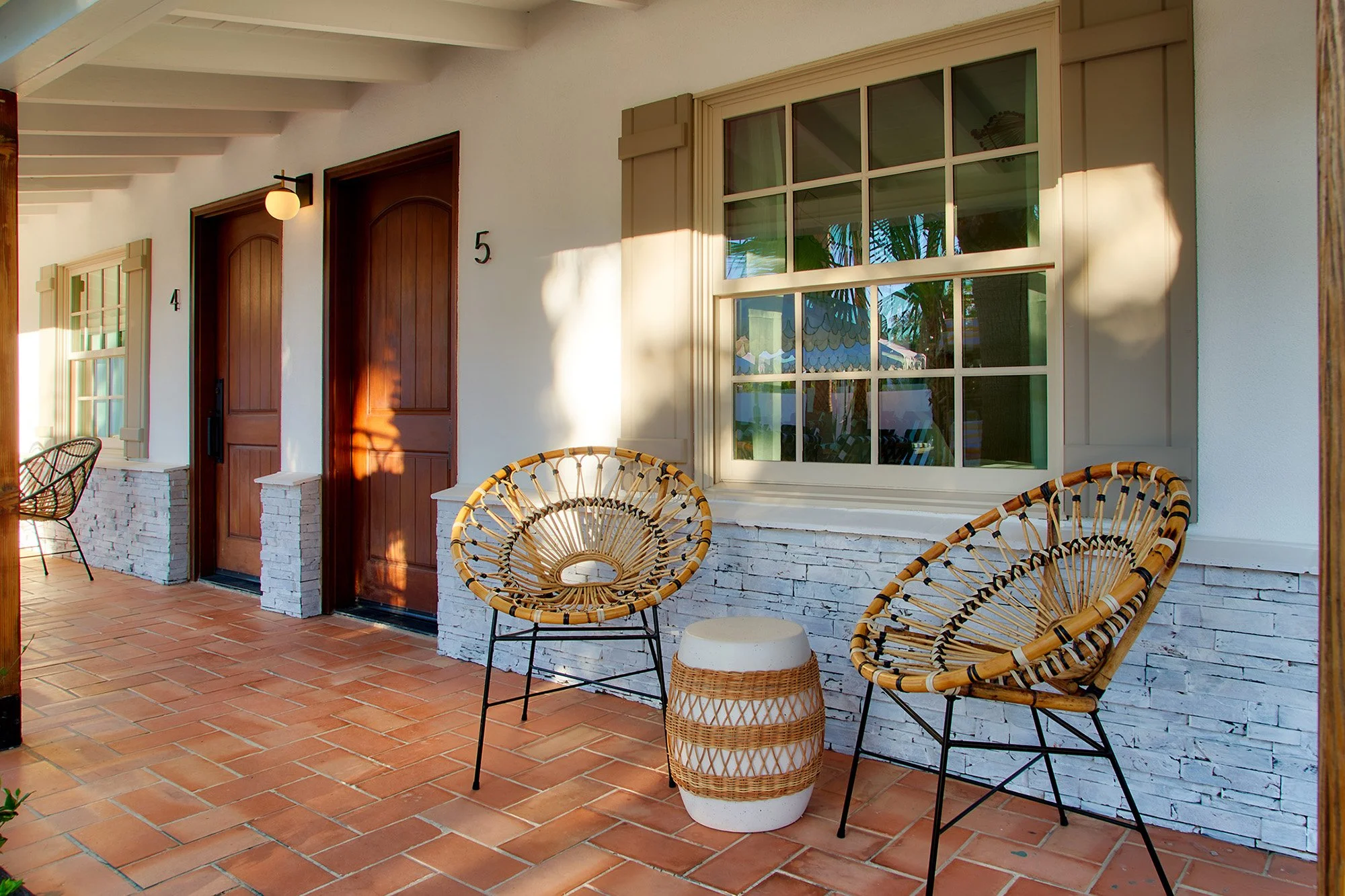 Cozy covered porch with two rattan chairs, a small side table, and a brick-floored seating area outside a white house with wood doors and large window. Sunlight casts shadows.