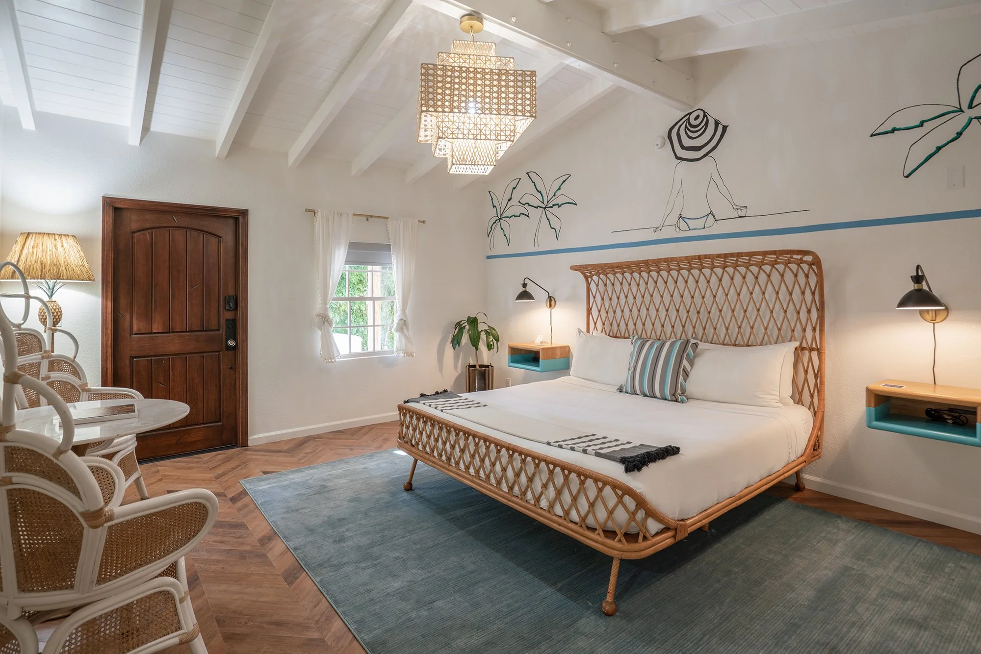 A bedroom with a wicker bed, wooden nightstands, and wall-mounted reading lamps. The room has white walls, a wooden door, a window with white curtains, and a wall mural of a woman in a hat with palm trees. There is a blue area rug on a wooden floor and a ceiling light fixture.