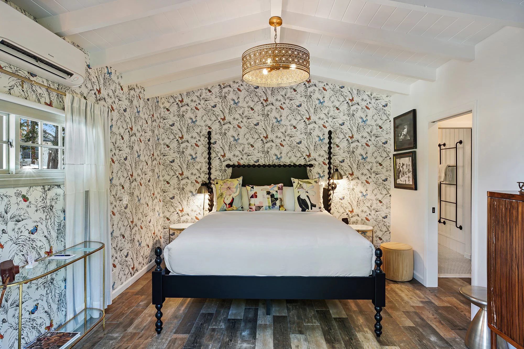 A bedroom with a four-poster bed against a wallpapered wall, featuring pillows with colorful animal illustrations, beside small bedside lamps, a window with white curtains, and a ceiling-mounted light fixture.