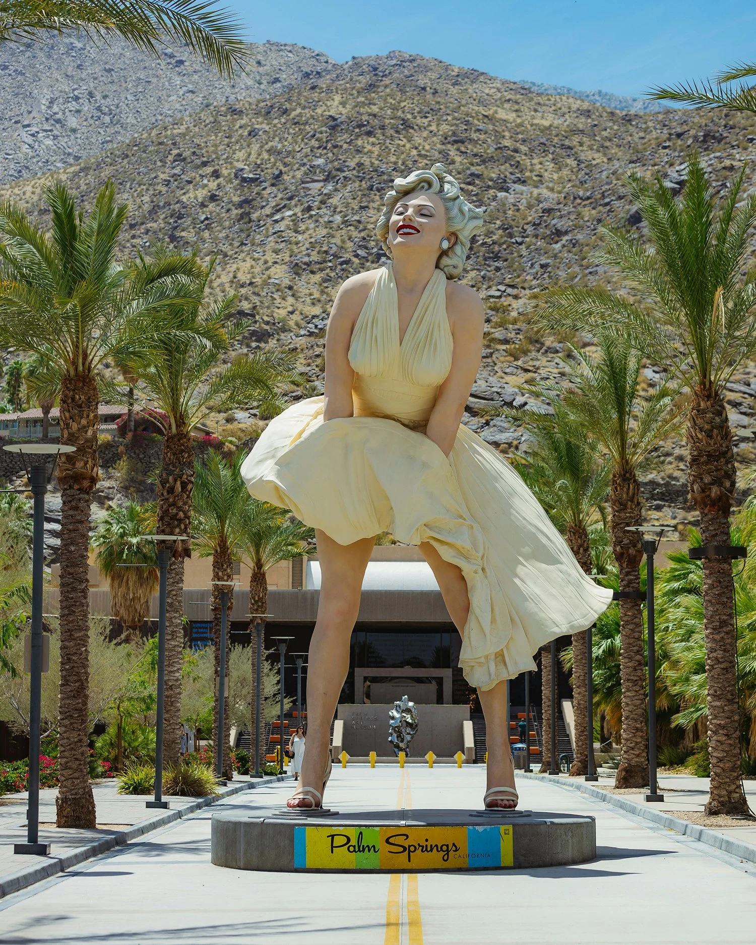 Large sculpture of Marilyn Monroe in a yellow dress, standing on a circular base with a 'Palm Springs California' sign, surrounded by palm trees in Palm Springs, California, with mountains in the background.