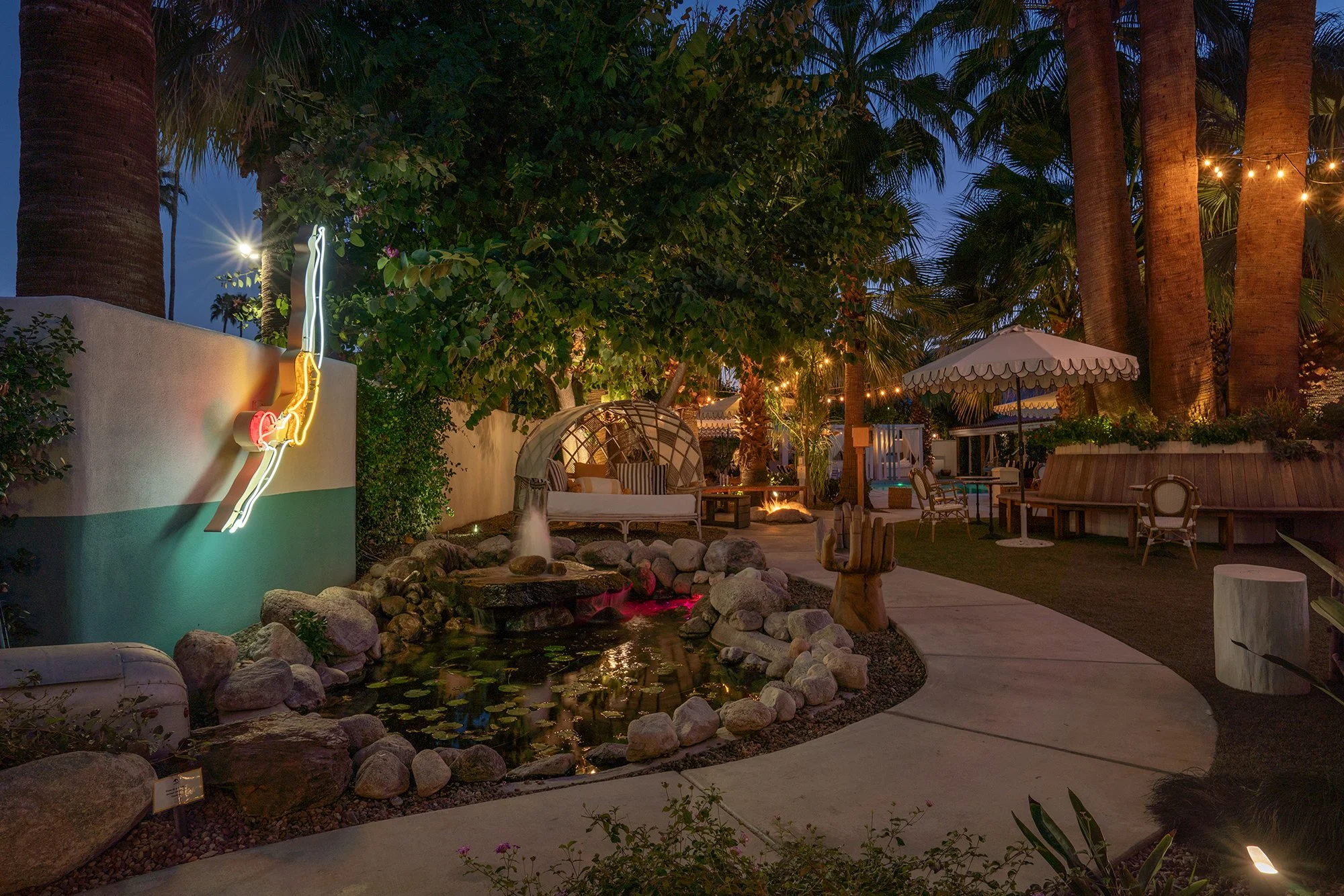 A cozy backyard at dusk decorated with string lights, featuring a pond with rocks, a fountain, outdoor seating including a swing, chairs, umbrellas, and lush greenery with tall palm trees.