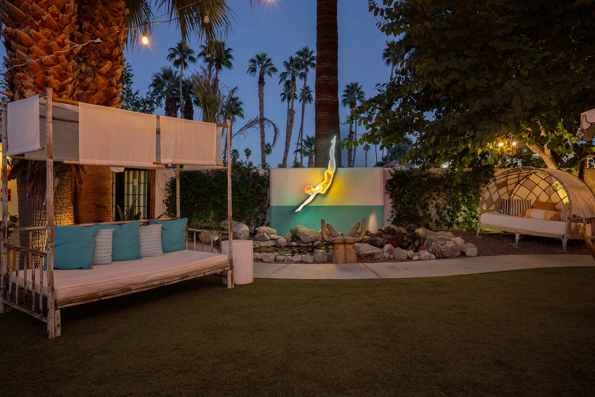 Outdoor patio with lounge chairs, a mural of a woman swimming, palm trees, and string lights at dusk.