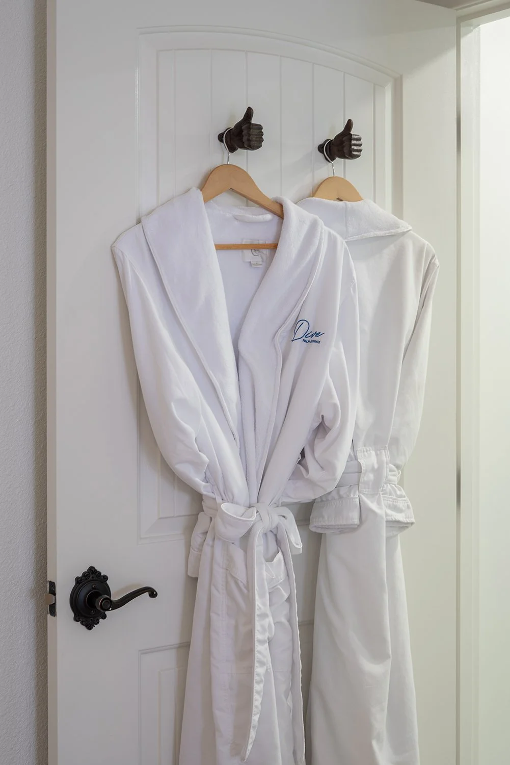 Two white bathrobes hanging on hooks shaped like hands giving a thumbs-up, attached to a white door.