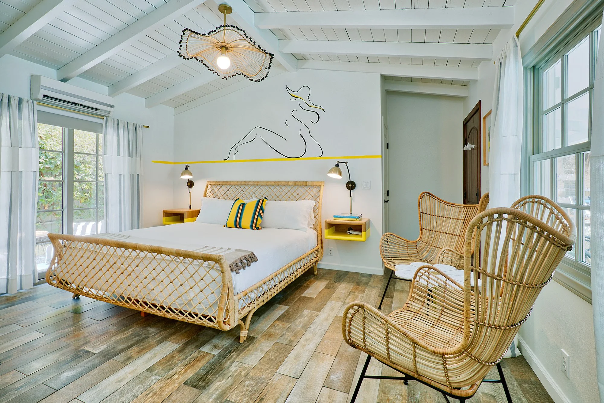 A bright bedroom with white walls, wooden flooring, and large windows with white curtains. It features a large bed with a rattan frame, white bedding, and colorful pillows. Two wall-mounted lamps are on either side of the bed. Above the bed, there's 