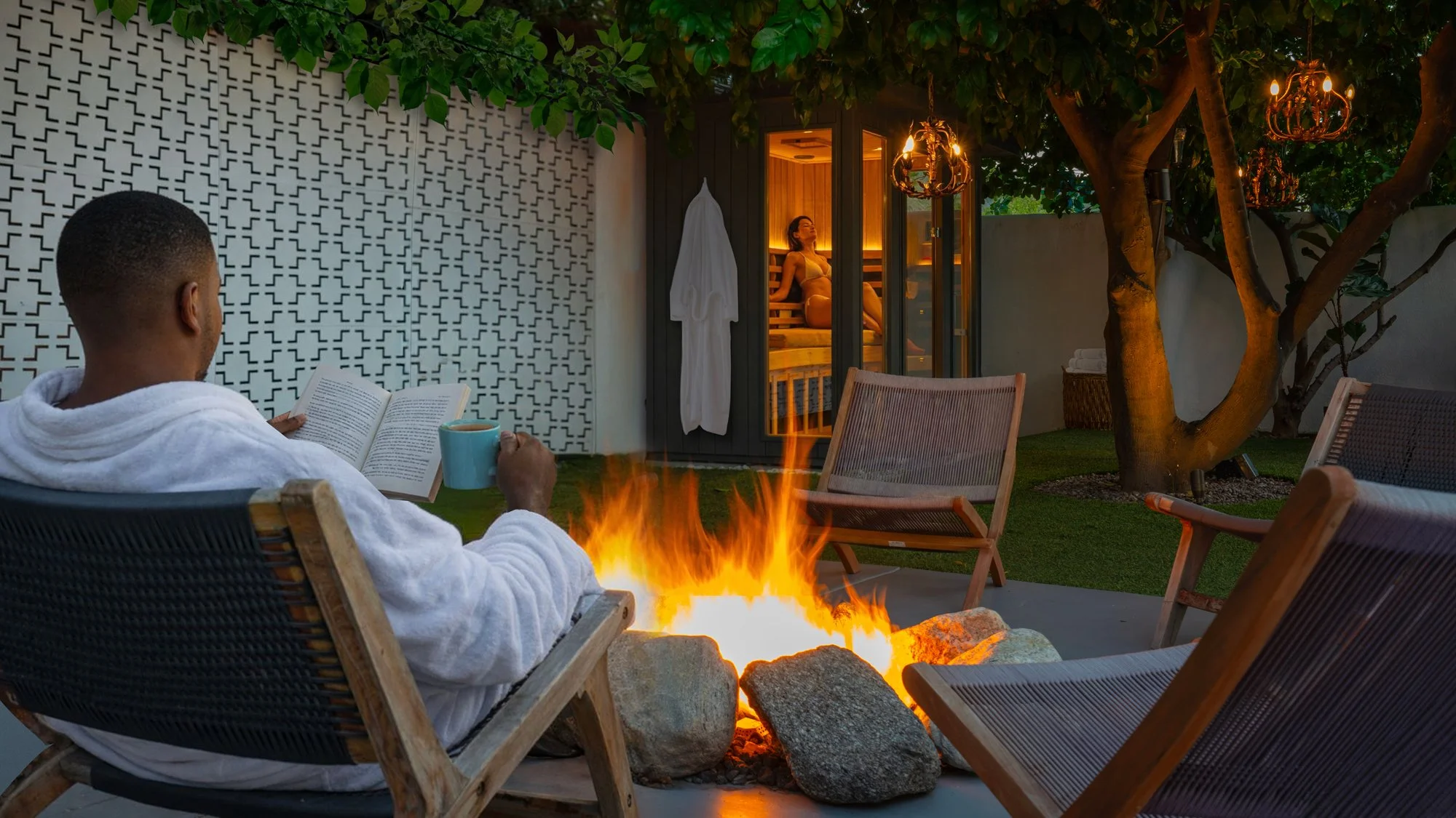 Person in a white robe sitting in outdoor chair, reading a book and holding a mug, next to a fire pit, with a woman in a sauna visible through glass doors in the background.