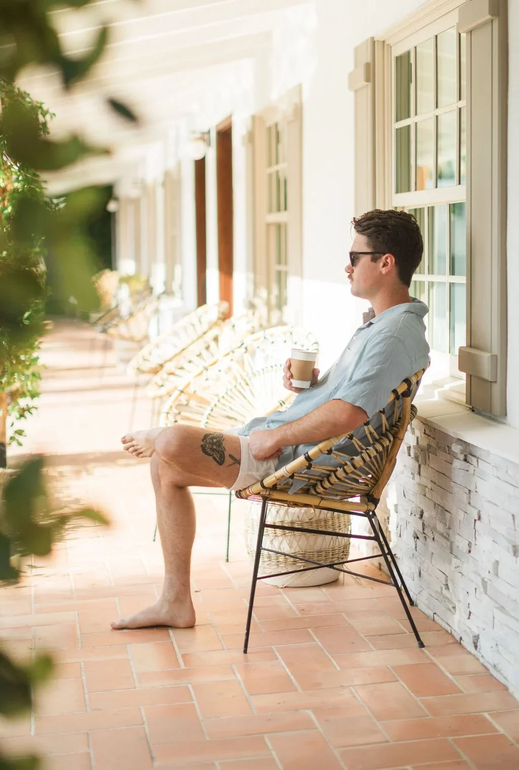 A man sitting on a wicker chair on a sunny patio, holding a coffee cup, wearing sunglasses with a tattoo on his thigh, dressed in a light blue shirt and beige shorts, next to a white brick wall and green plants.