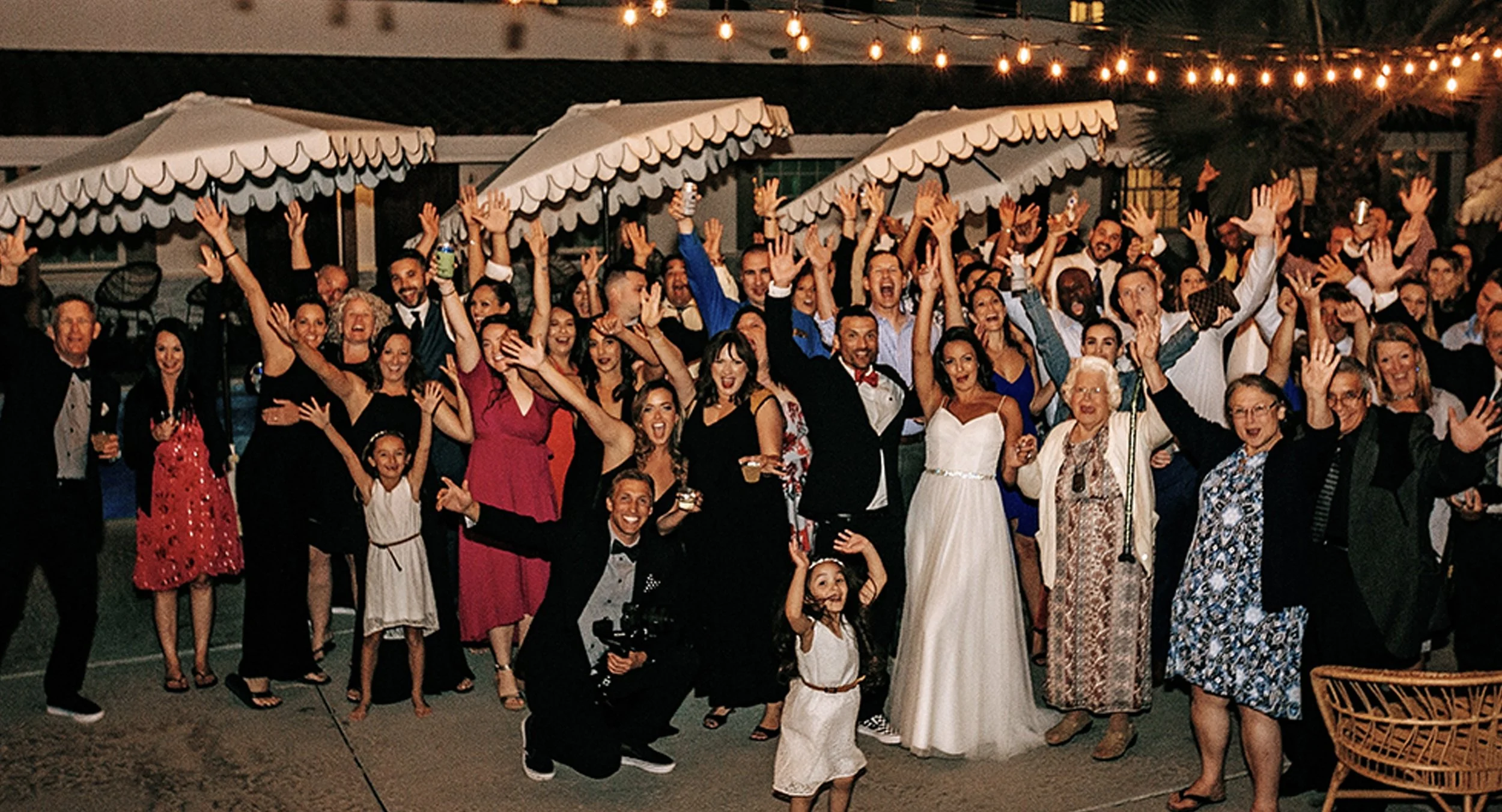 Group of people celebrating at a party or wedding reception, smiling, raising hands, and wearing formal attire, with string lights overhead.