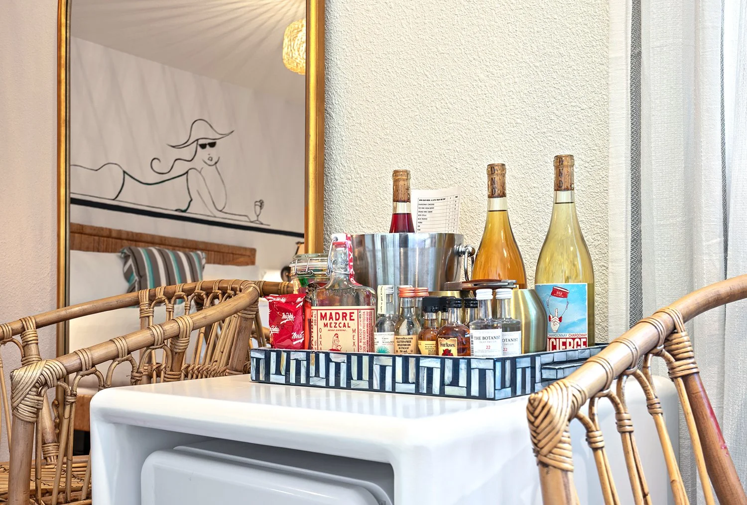 A tray on a table holding liquor bottles, wine bottles, and small bottles, with a metal ice bucket, in a room with wicker chairs and a wall mirror.