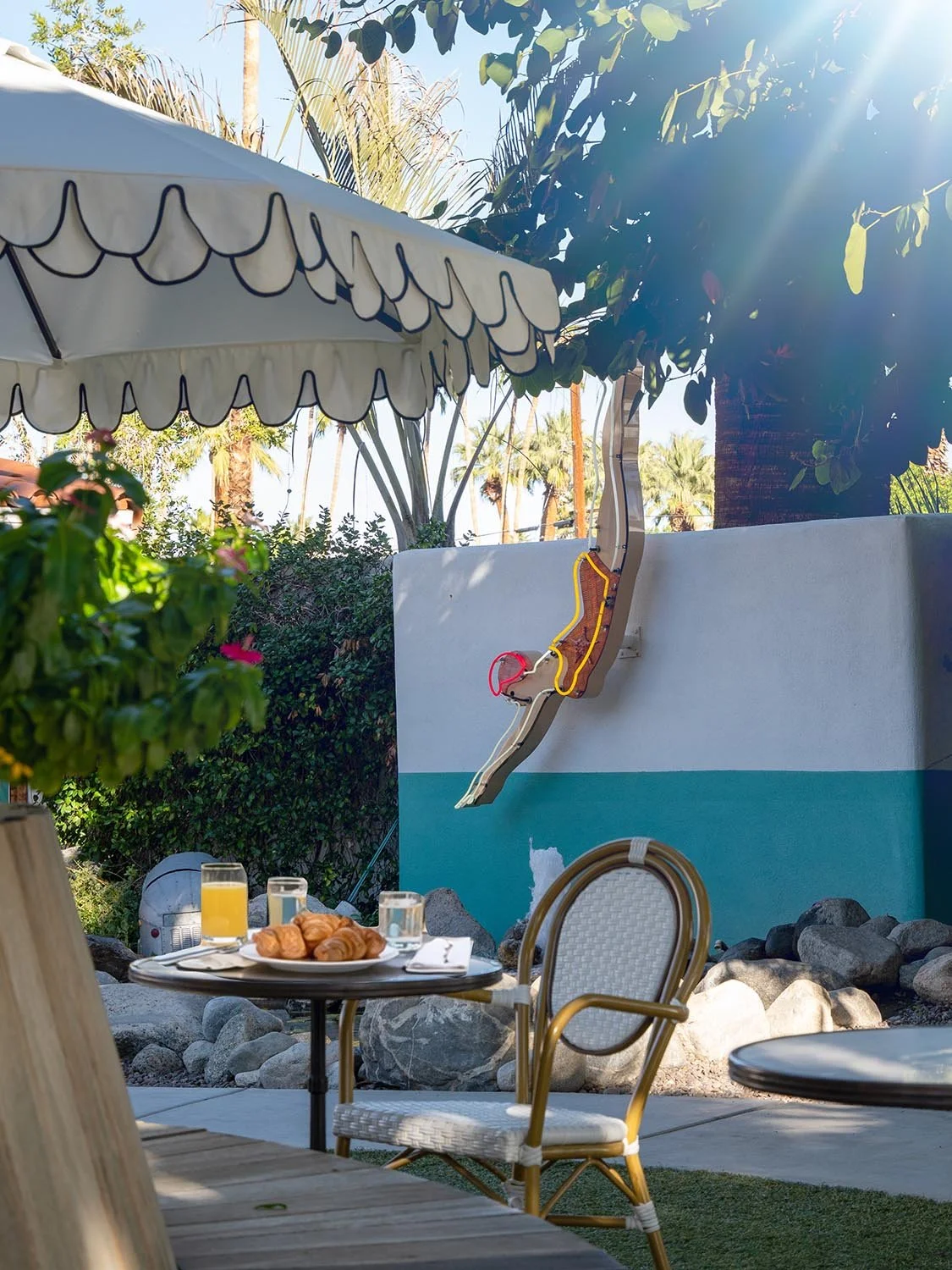 Outdoor patio scene with a table holding orange juice and pastries, surrounded by chairs, with a partly cloudy sky, palm trees, and a whimsical water slide shaped like a figure in the background.