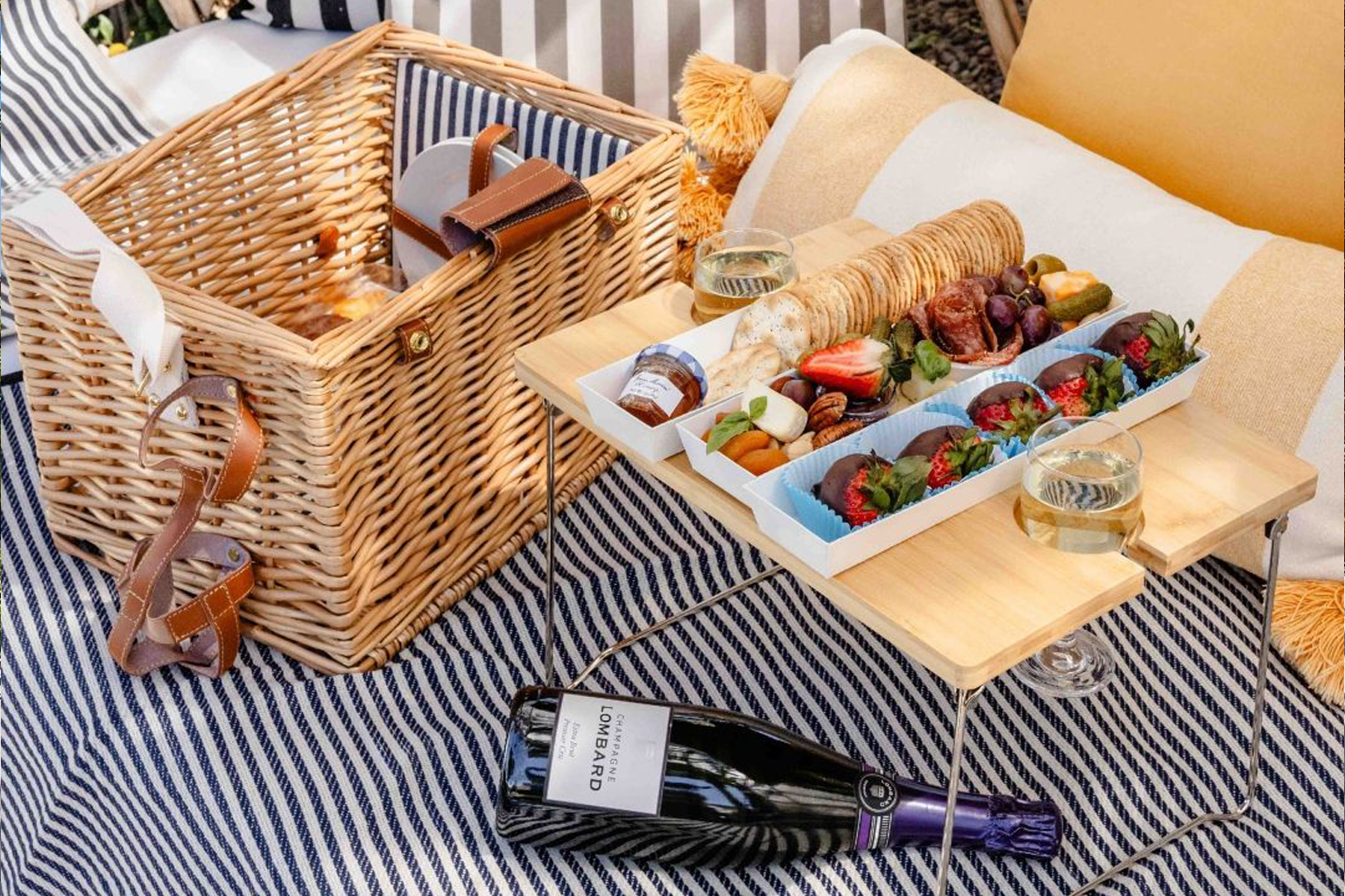 Picnic setup with a wicker basket, a wooden tray with assorted snacks, strawberries, and glasses of white wine on a striped tablecloth.