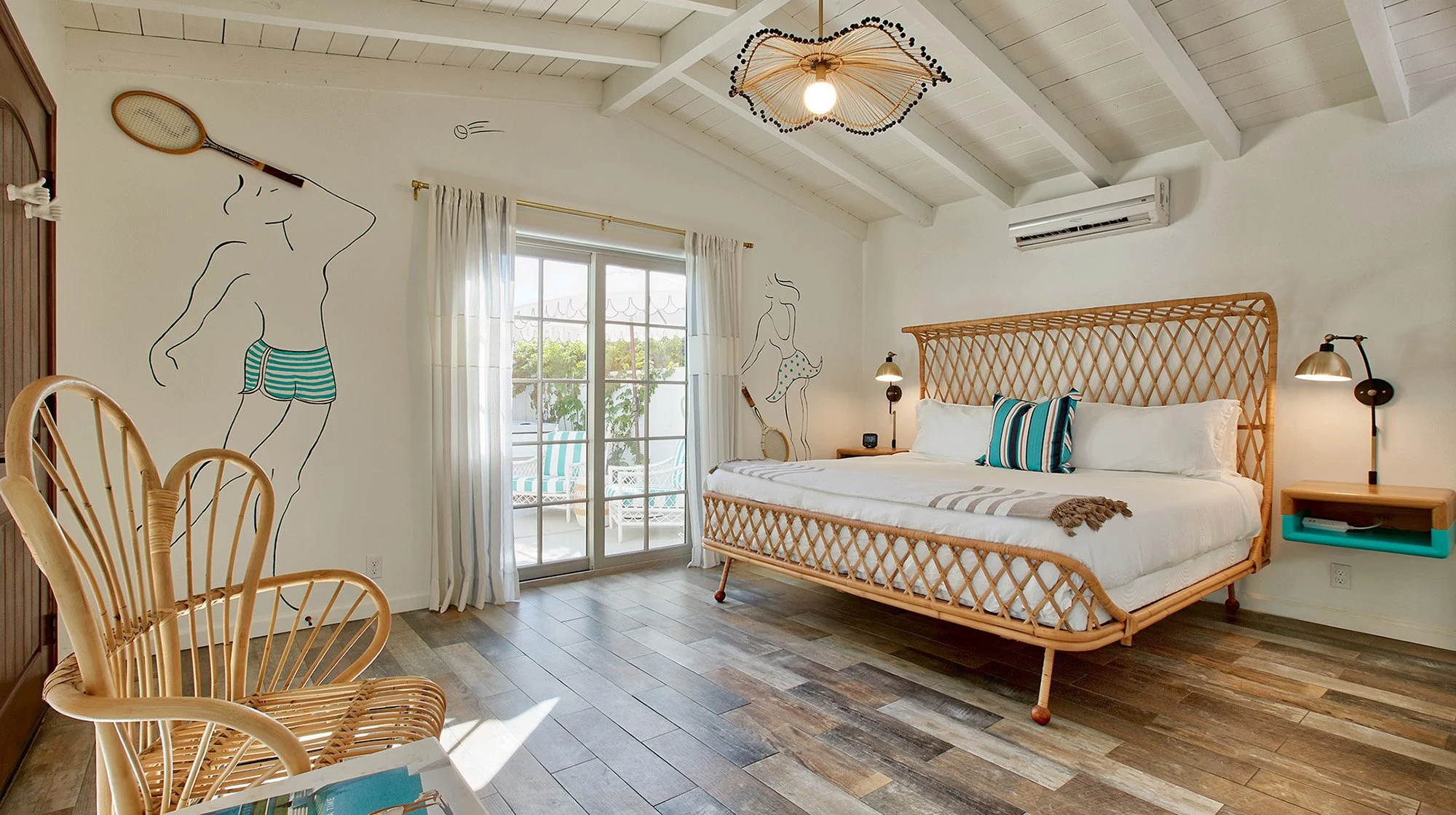 Bright beach-themed bedroom with a rattan bed, striped pillows, and wall art of a woman in swimwear; sliding glass door leads to an outdoor patio.