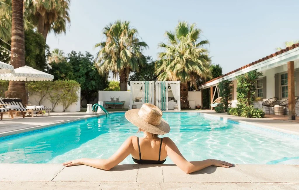 A woman sitting at the edge of a swimming pool, wearing a large straw hat and a black swimsuit, surrounded by palm trees and white outdoor furniture.