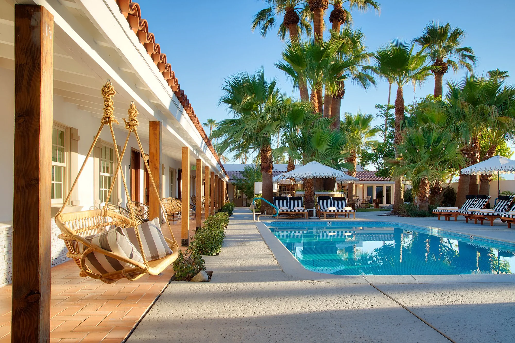 A luxurious outdoor pool area with lounge chairs, umbrellas, palm trees, and a building with hammock swings.