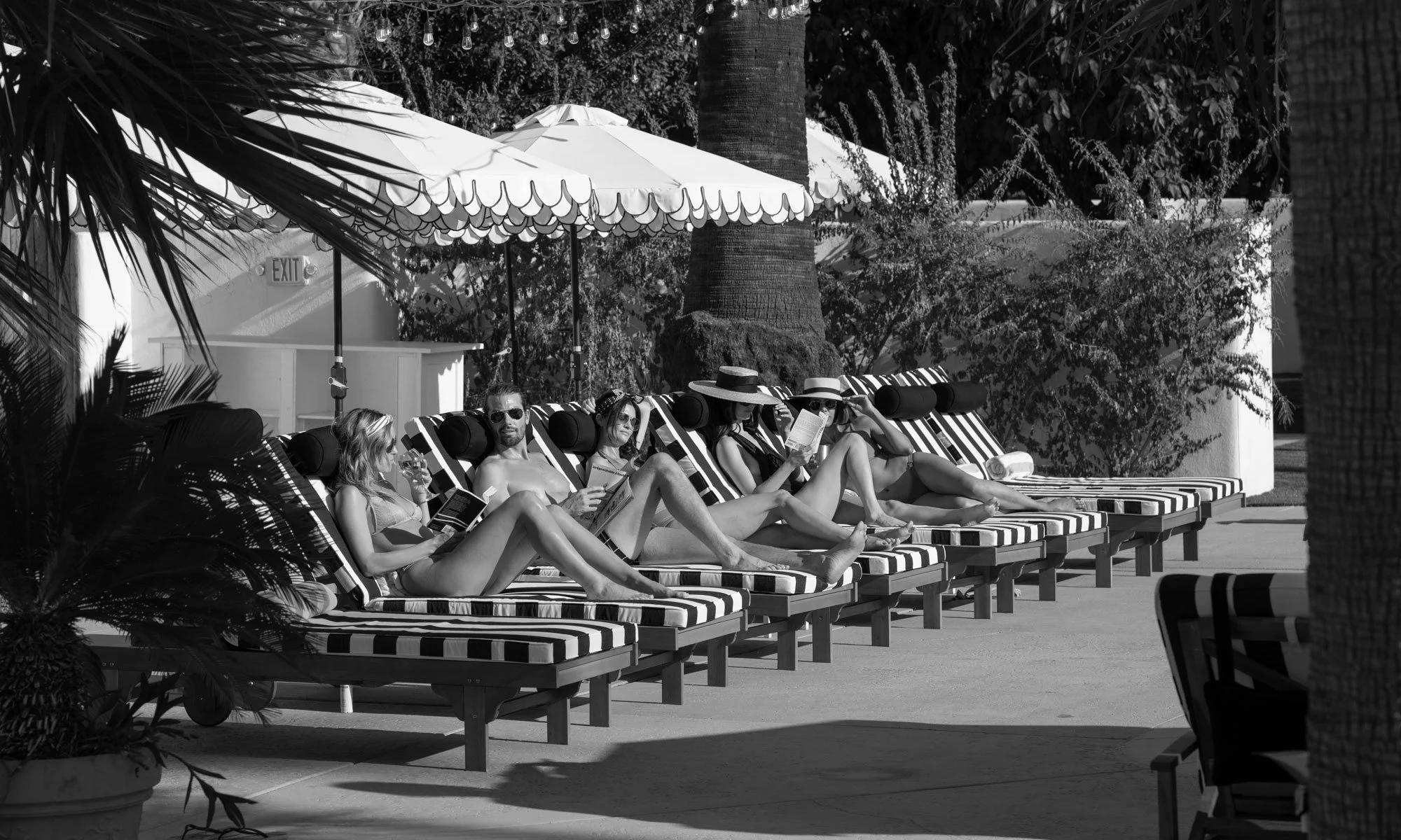A black and white photo of five people relaxing on striped lounge chairs by a pool under umbrellas, some reading and some talking, with trees and plants in the background.
