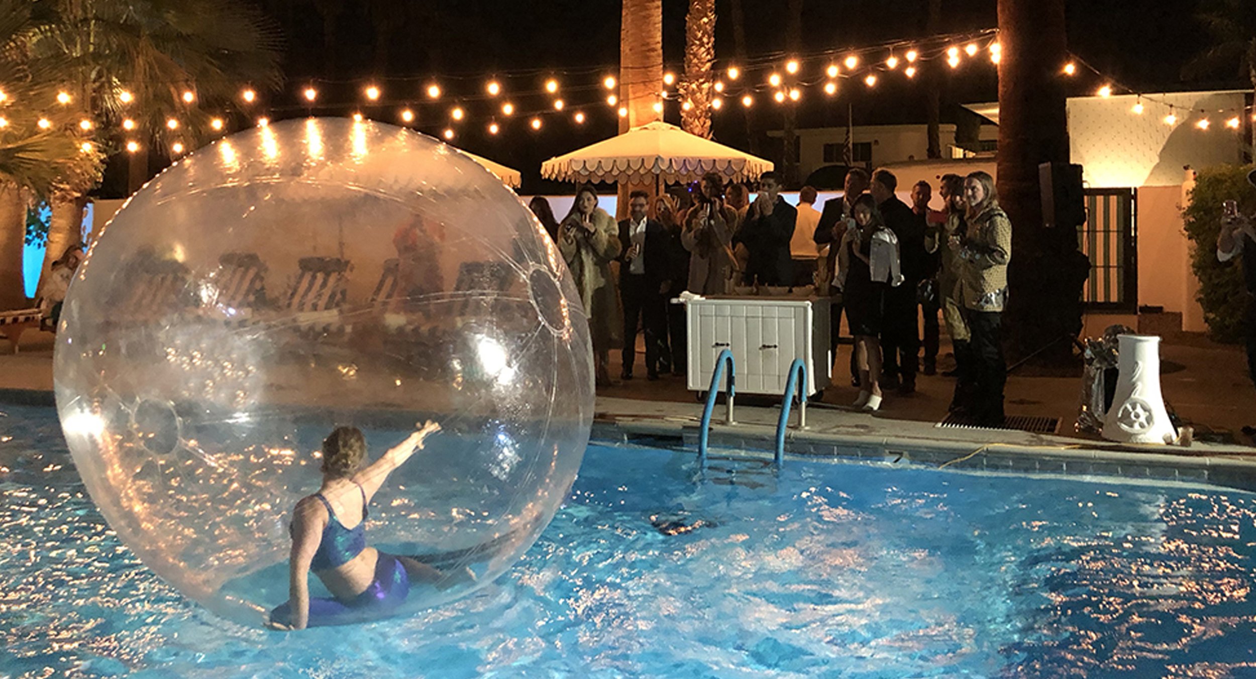 Person inside a large transparent inflatable ball floating in a swimming pool at night, with a crowd of people gathered nearby under string lights and outdoor umbrellas.
