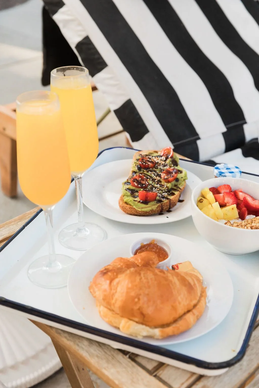 A tray with two glasses of mimosa, a slice of avocado toast topped with cherry tomatoes and sesame seeds, a bowl of fruit with strawberries and pineapple, a croissant with jam, and a small dish of preserve on a table with a striped black and white um