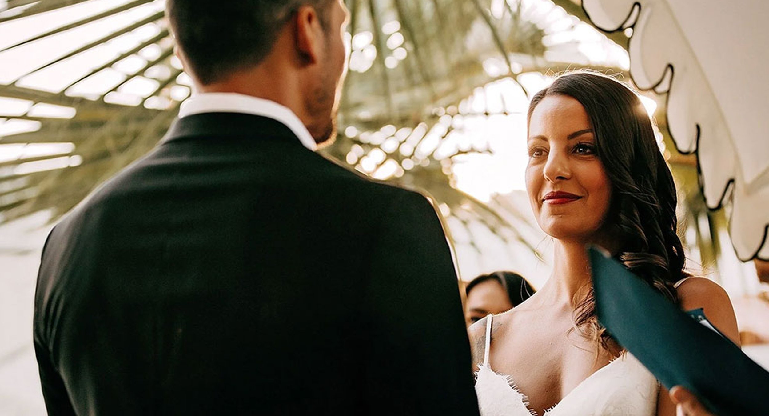 A bride and groom exchanging vows during their wedding ceremony under a canopy of tropical palm leaves.