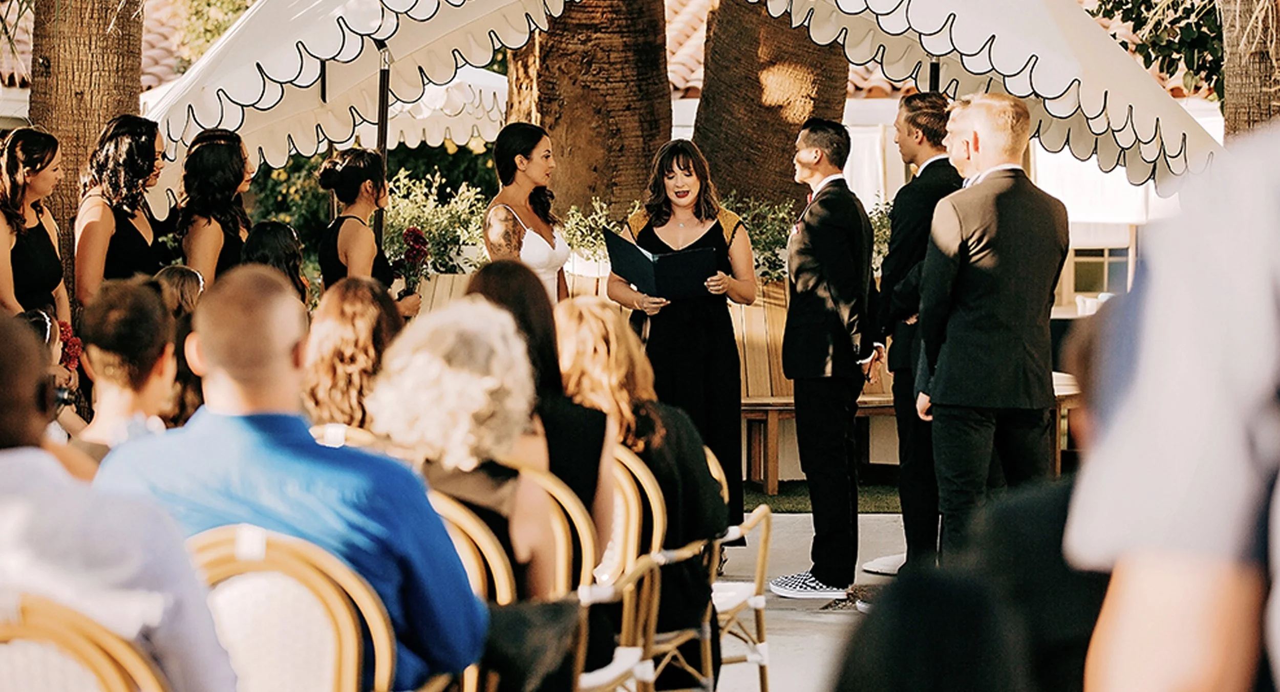 A wedding ceremony outdoors with a bride, groom, and officiant, surrounded by guests and wedding party under a canopy with floral decorations.