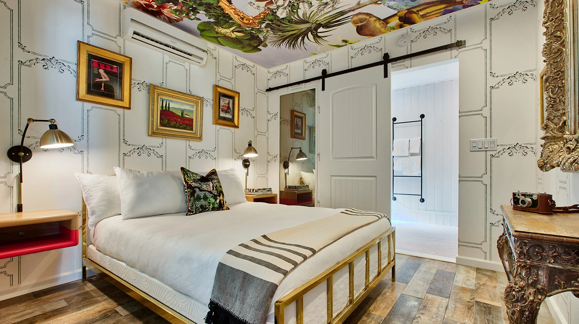 A bedroom with a white bed, decorative pillows, two wall-mounted lamps, framed artwork on the walls, an ornate mirror, wooden floors, an air conditioner, and a doorway leading to another room with white paneling.