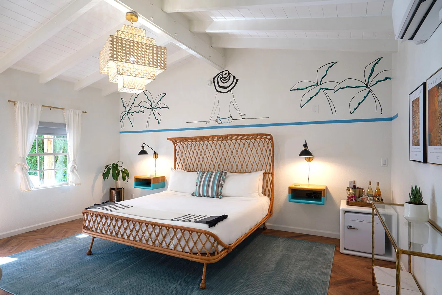 Bedroom with white walls, wooden floor, and a large bed with a rattan headboard. Above the bed, there is wall art of a woman in a bikini with a hat and palm trees. The room has a window with white curtains, a blue rug, and two black wall lamps. There