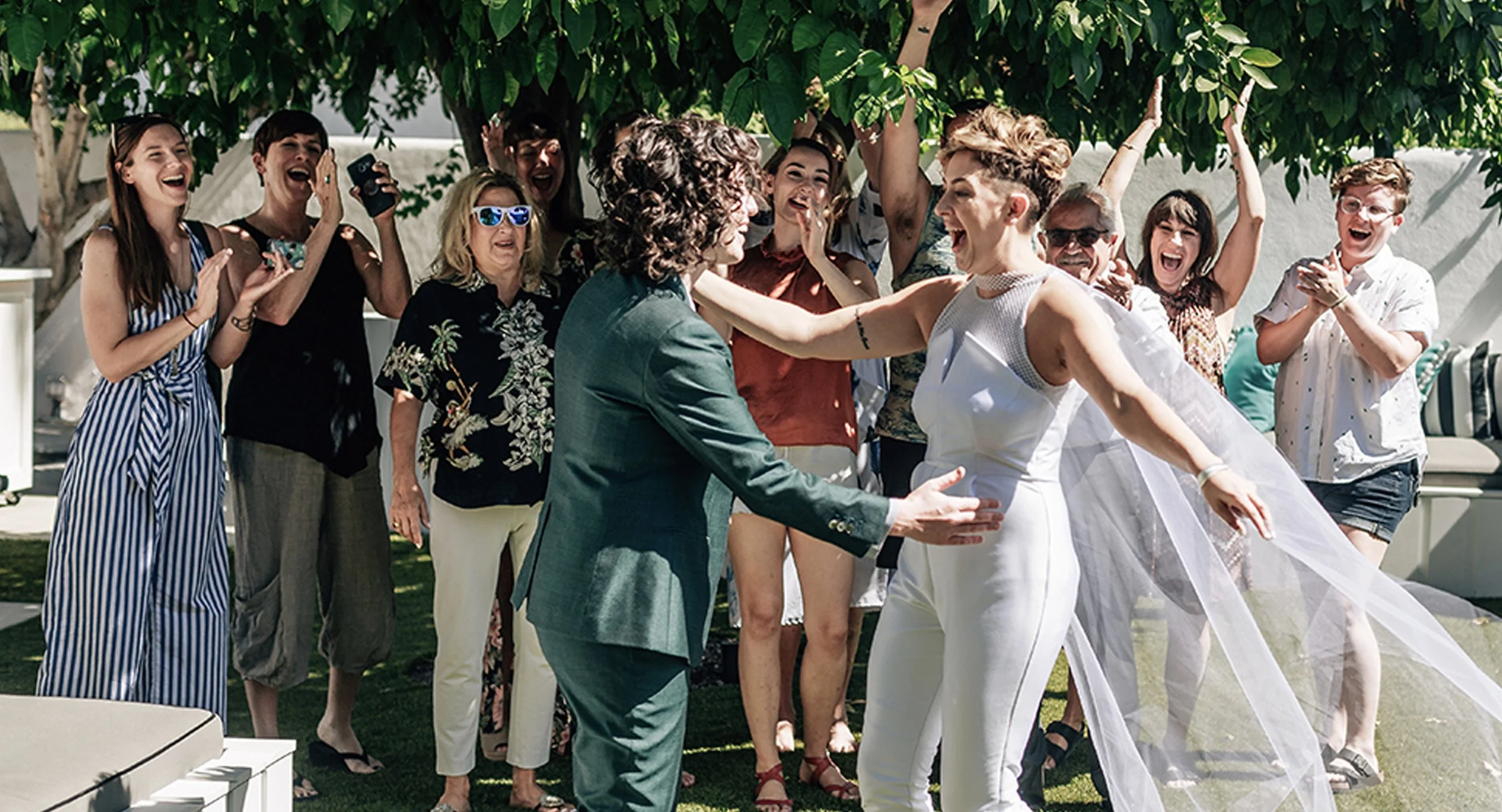 A group of people at a wedding celebration outdoors, with two women in the foreground dancing and smiling, surrounded by friends and family who are clapping and cheering.