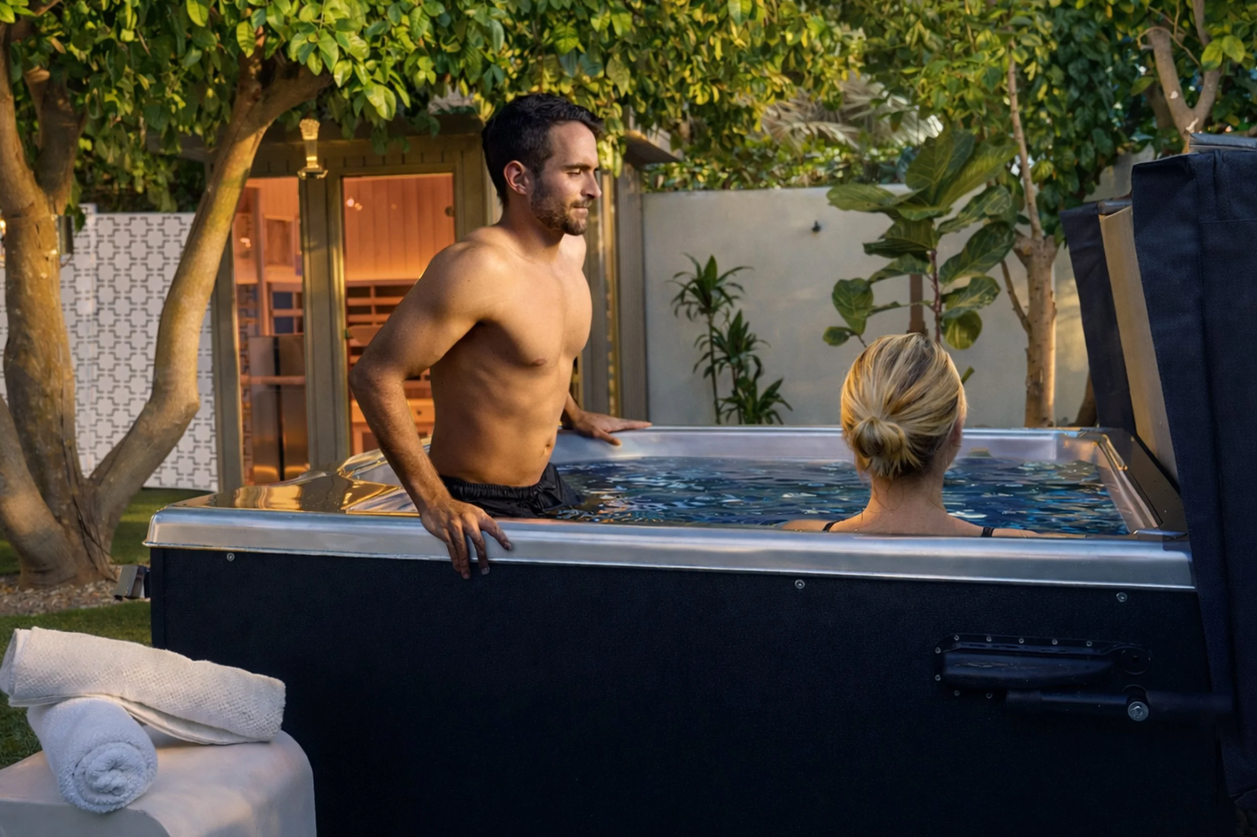 A man and woman in an outdoor hot tub surrounded by trees and plants in a backyard at dusk.