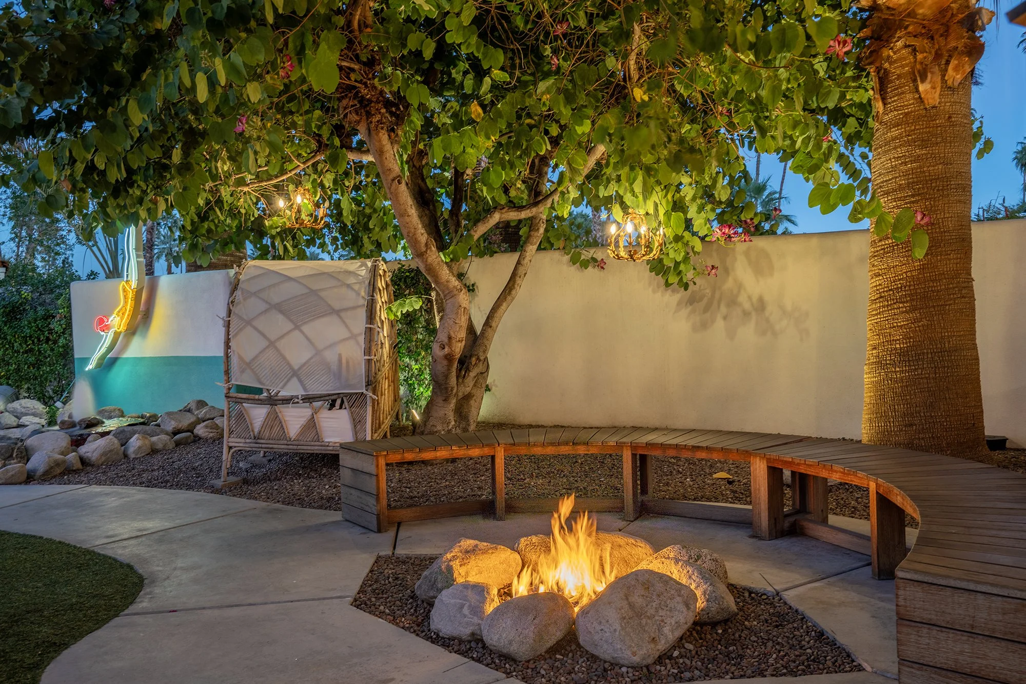 Backyard patio at dusk with a fire pit surrounded by rocks, a curved wooden bench, a tree with hanging lantern-style lights, a white wall with neon art, and a tall palm tree.