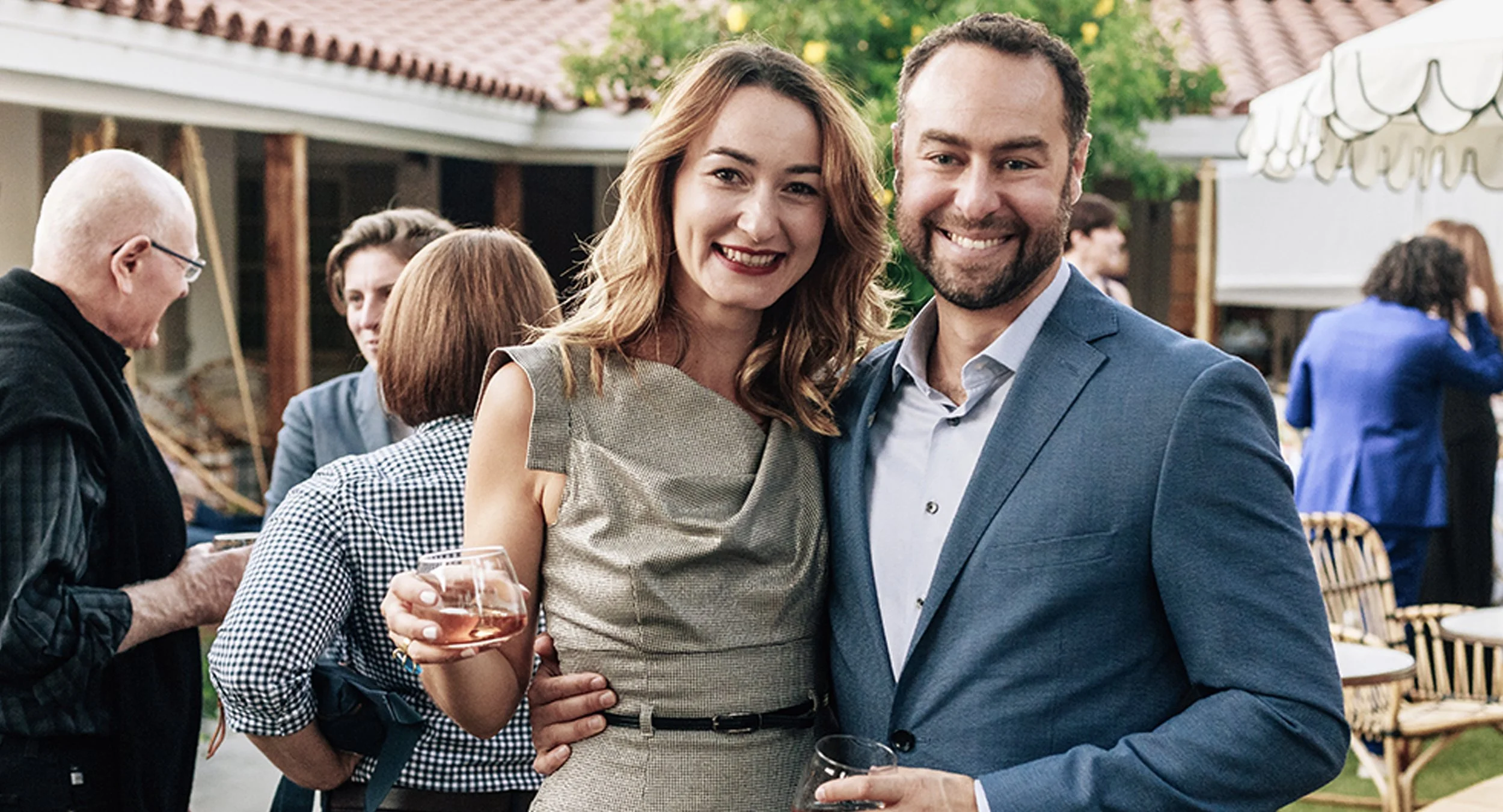 Couple smiling and holding drinks at an outdoor social gathering with other people in the background.
