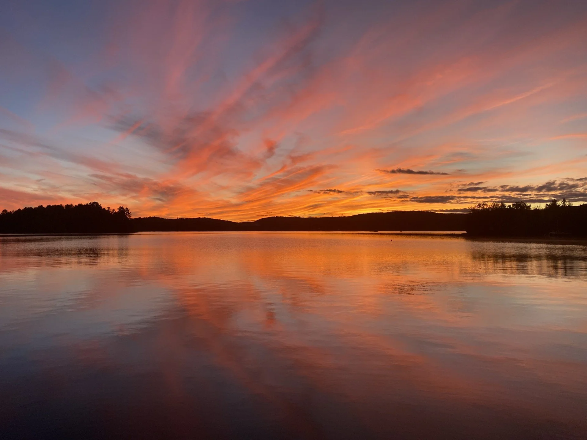 A colorful sunset with orange, pink, and purple clouds over a calm lake, with silhouettes of trees and hills in the distance.