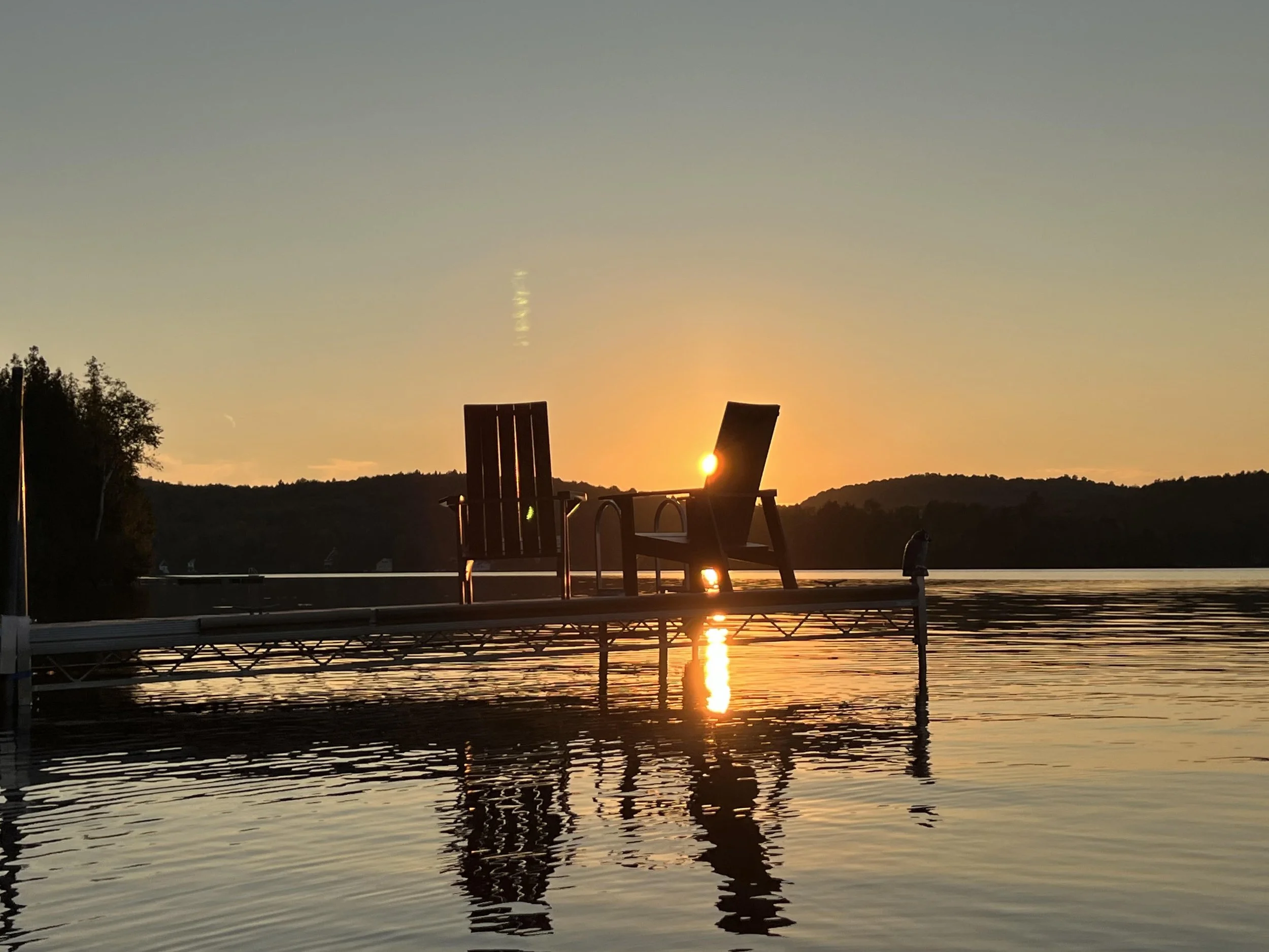 Silhouette of two Adirondack chairs on a dock at sunset over a lake, with hills in the background and calm water reflecting the sky's warm colors.