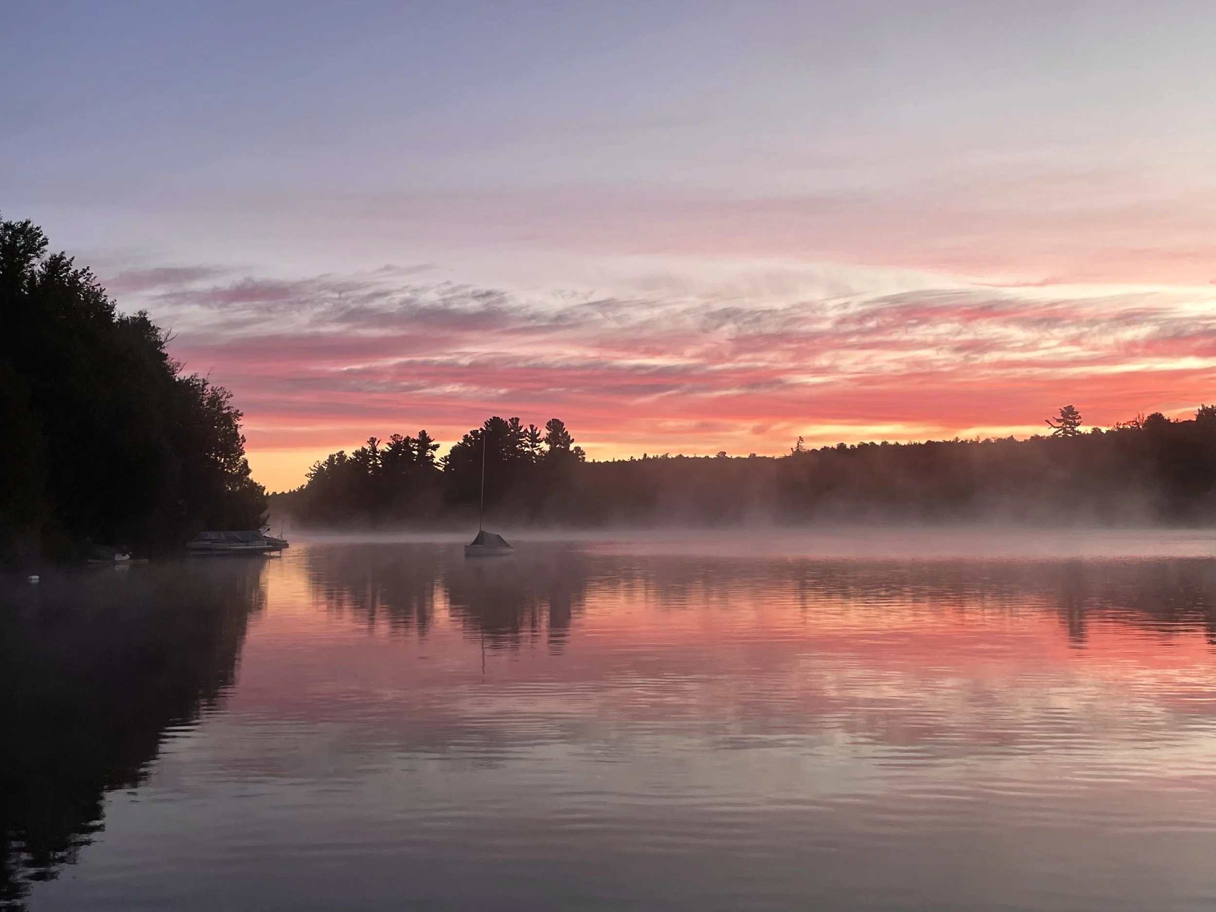 A calm body of water at sunrise with colorful pink, orange, and purple clouds, surrounded by trees and mist on the water.