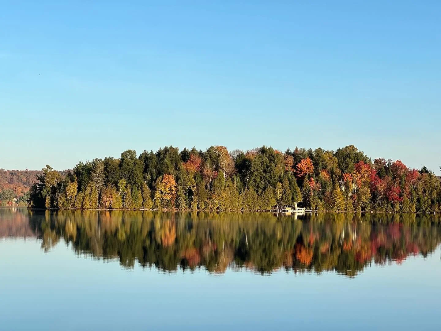 A calm lake reflecting a forested shoreline with autumn trees and a clear blue sky.
