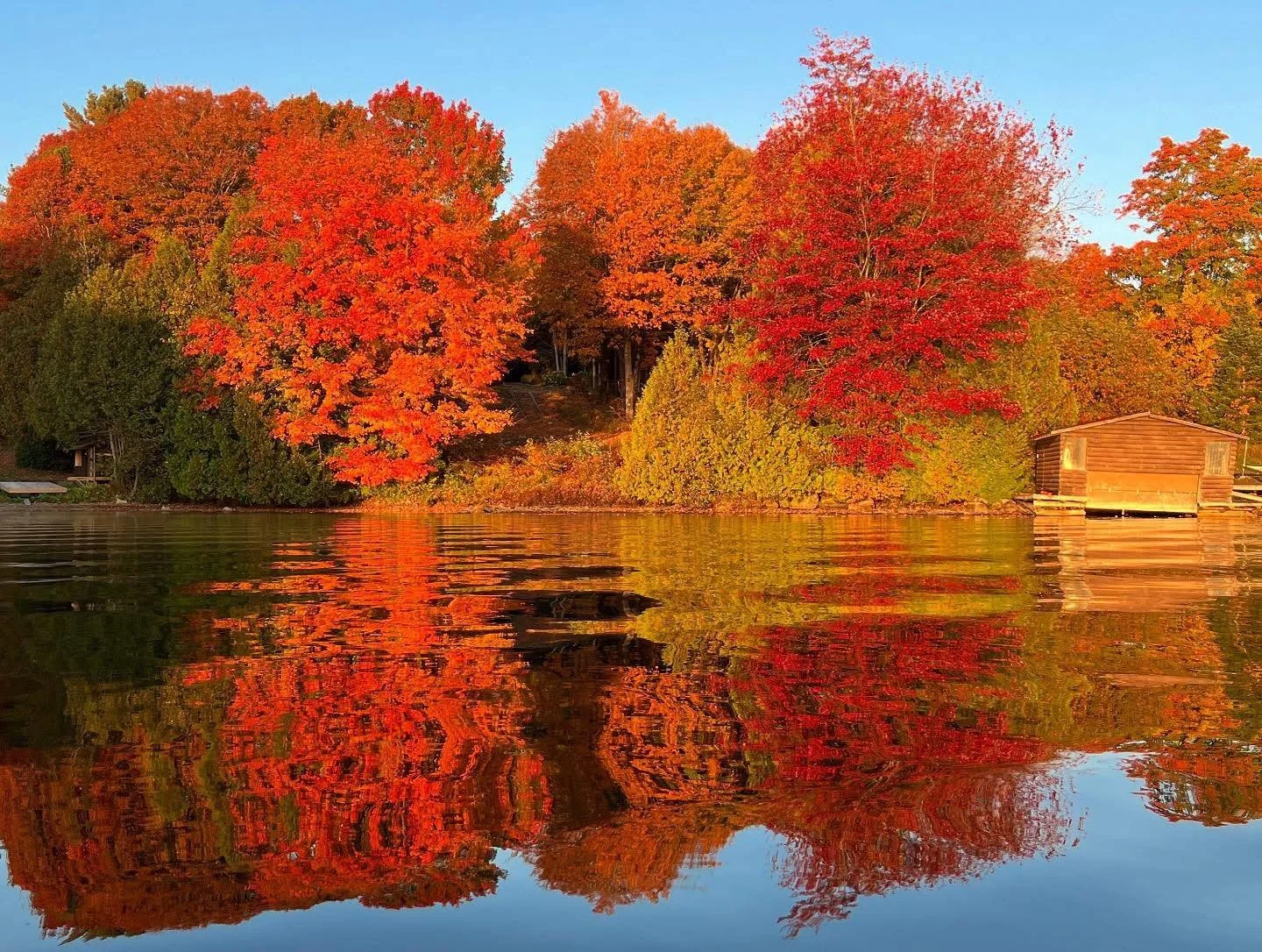Autumn scene with colorful orange and red trees reflected in a lake, and a small wooden boathouse on the right side.