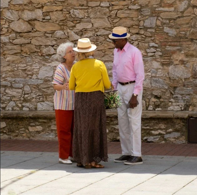 Three elderly people, two women and one man, standing outdoors against a stone wall talking casually. The woman on the left has white curly hair, wears a colorful striped shirt and red pants. The woman in the middle has blonde hair, a yellow top, and a long patterned skirt, holding a green patterned bag. The man on the right wears a pink shirt, white pants, and a straw hat with a blue band.