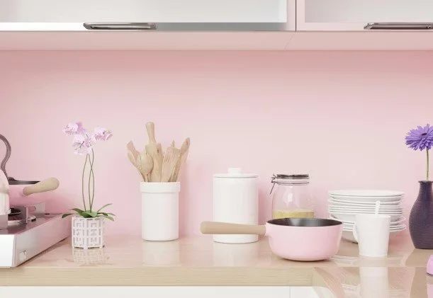 Kitchen countertop with pink and purple flowers, a white container holding wooden utensils, a pink pot, plates, and jars on a pastel pink wall background.