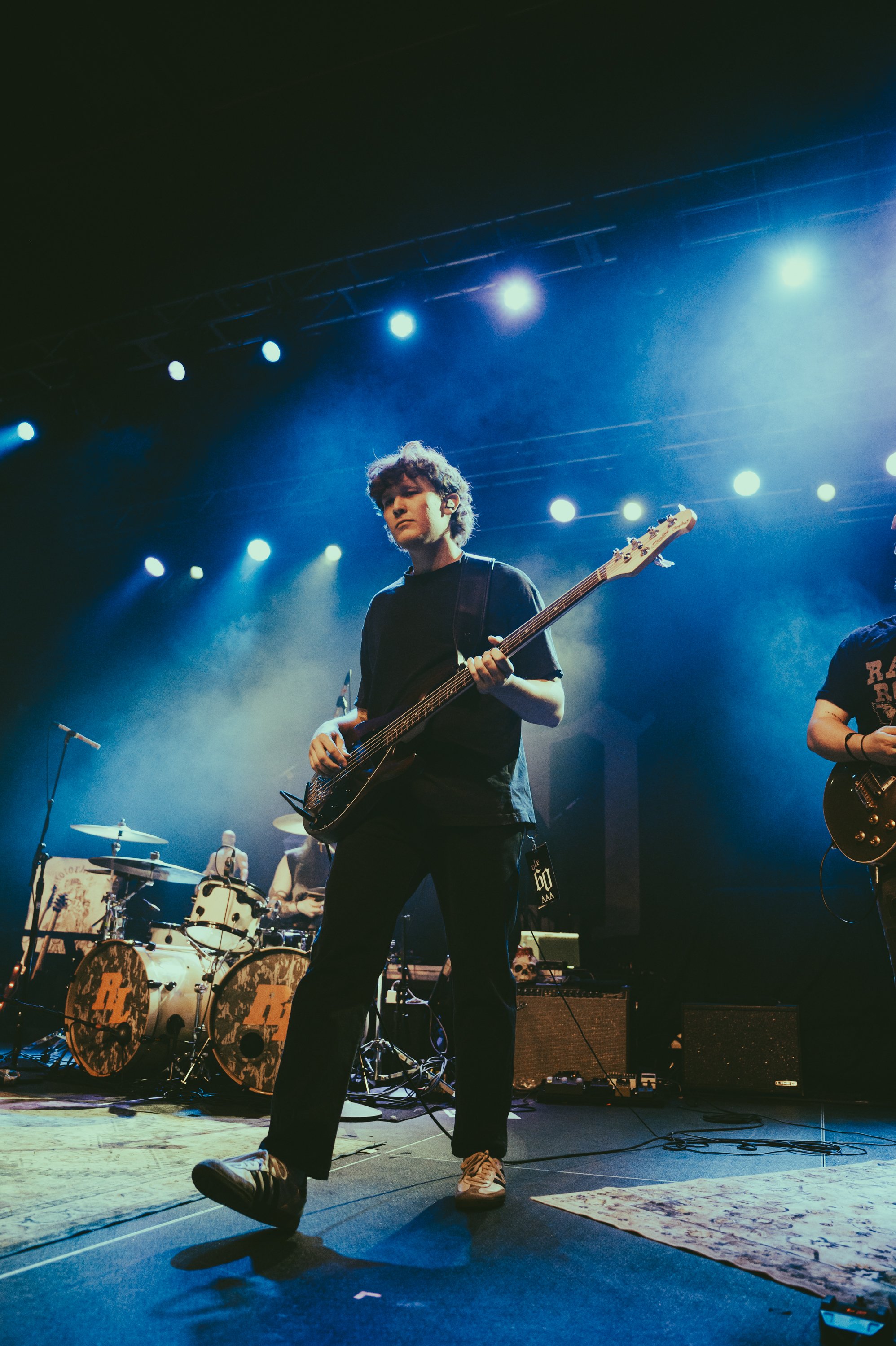 A young musician playing a bass guitar on stage during a concert, with stage lights shining and a drummer visible in the background.