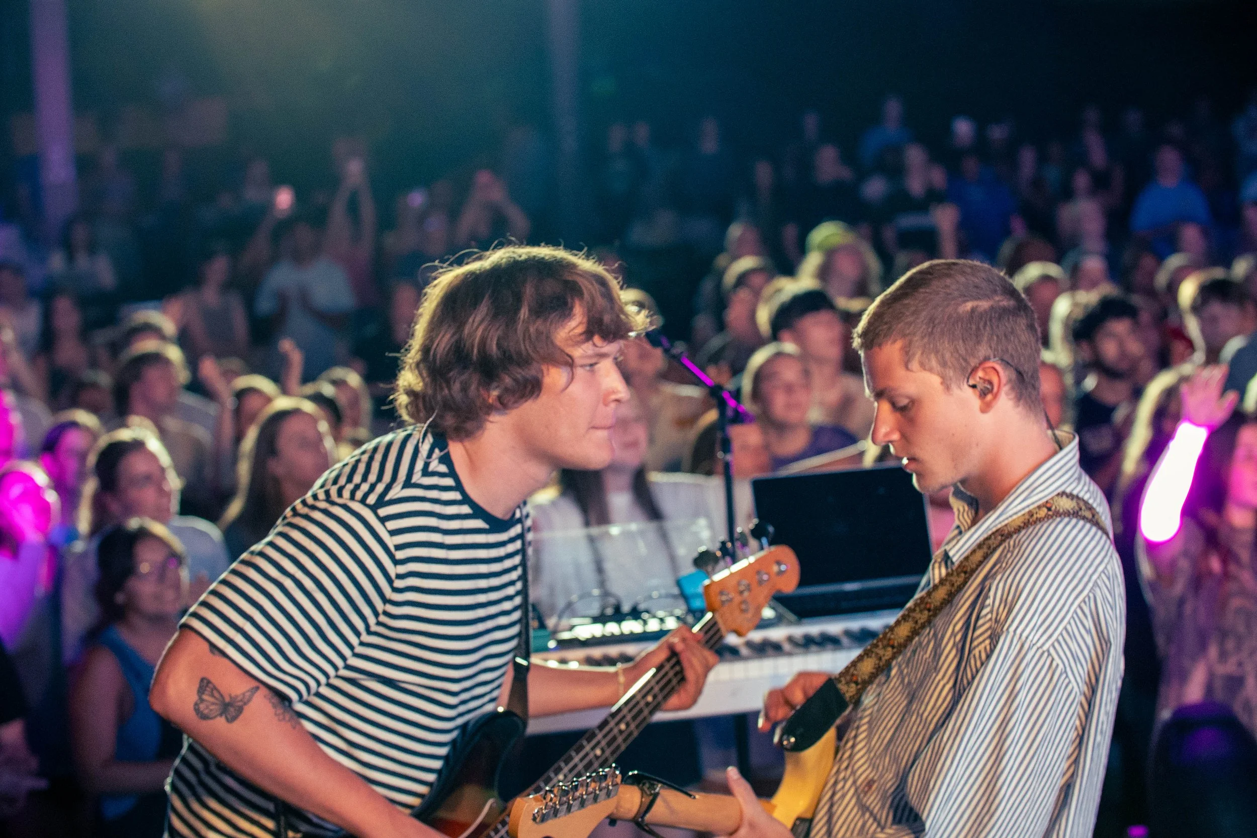 Two young men performing music together in front of an audience. One is playing an acoustic guitar and the other is playing a keyboard, both focused on their instruments. The audience is watching and clapping, with colorful stage lighting illuminating the scene.