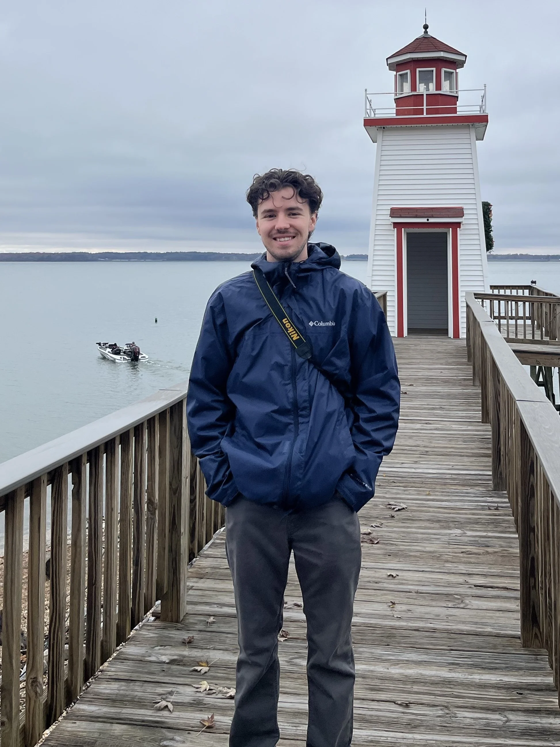 A young man in a blue jacket and gray pants standing on a wooden dock in front of a white lighthouse with red accents, near a body of water with a boat and distant shoreline under an overcast sky.