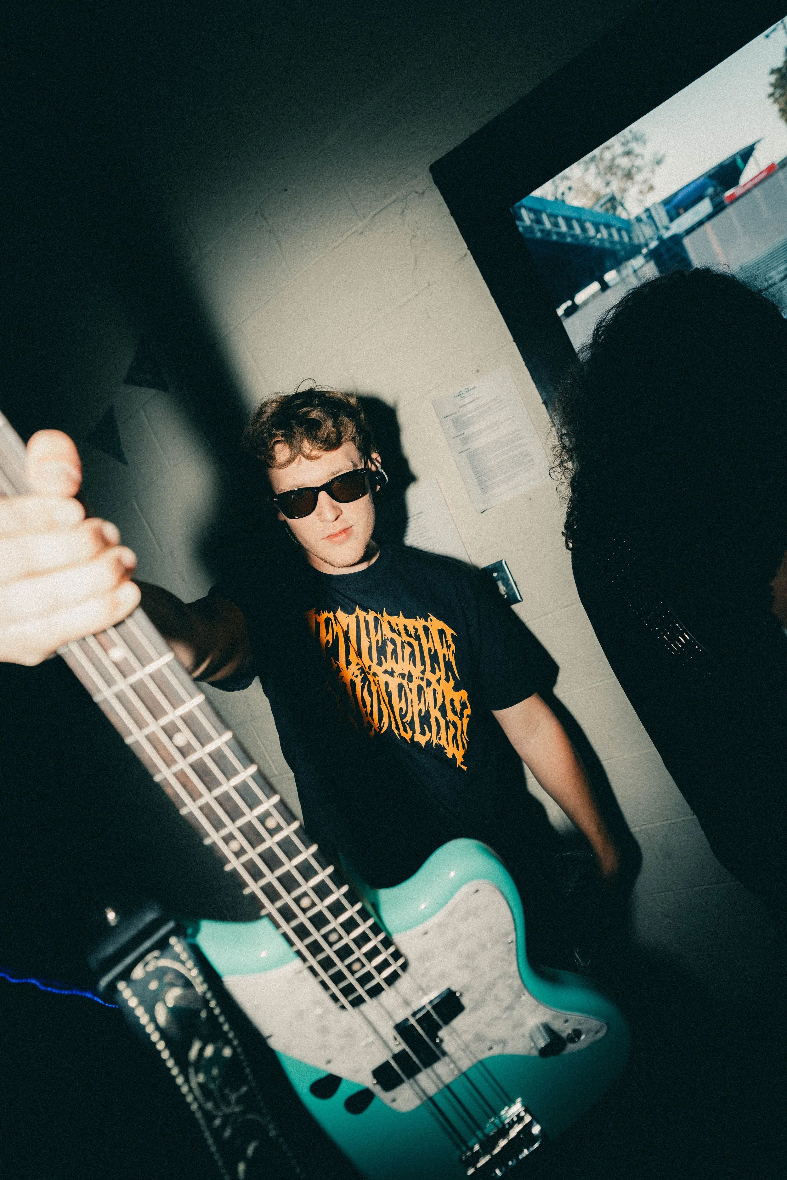 Young man with sunglasses and a black T-shirt playing an electric guitar, standing inside a room with a window showing outdoor equipment or construction in the background.