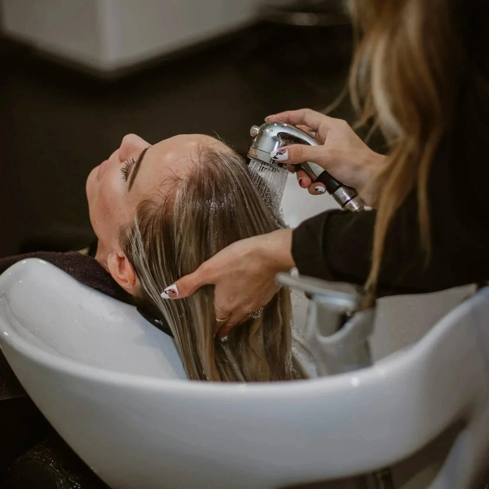 A woman lying back in a salon sink having her hair washed by a stylist using a handheld showerhead.